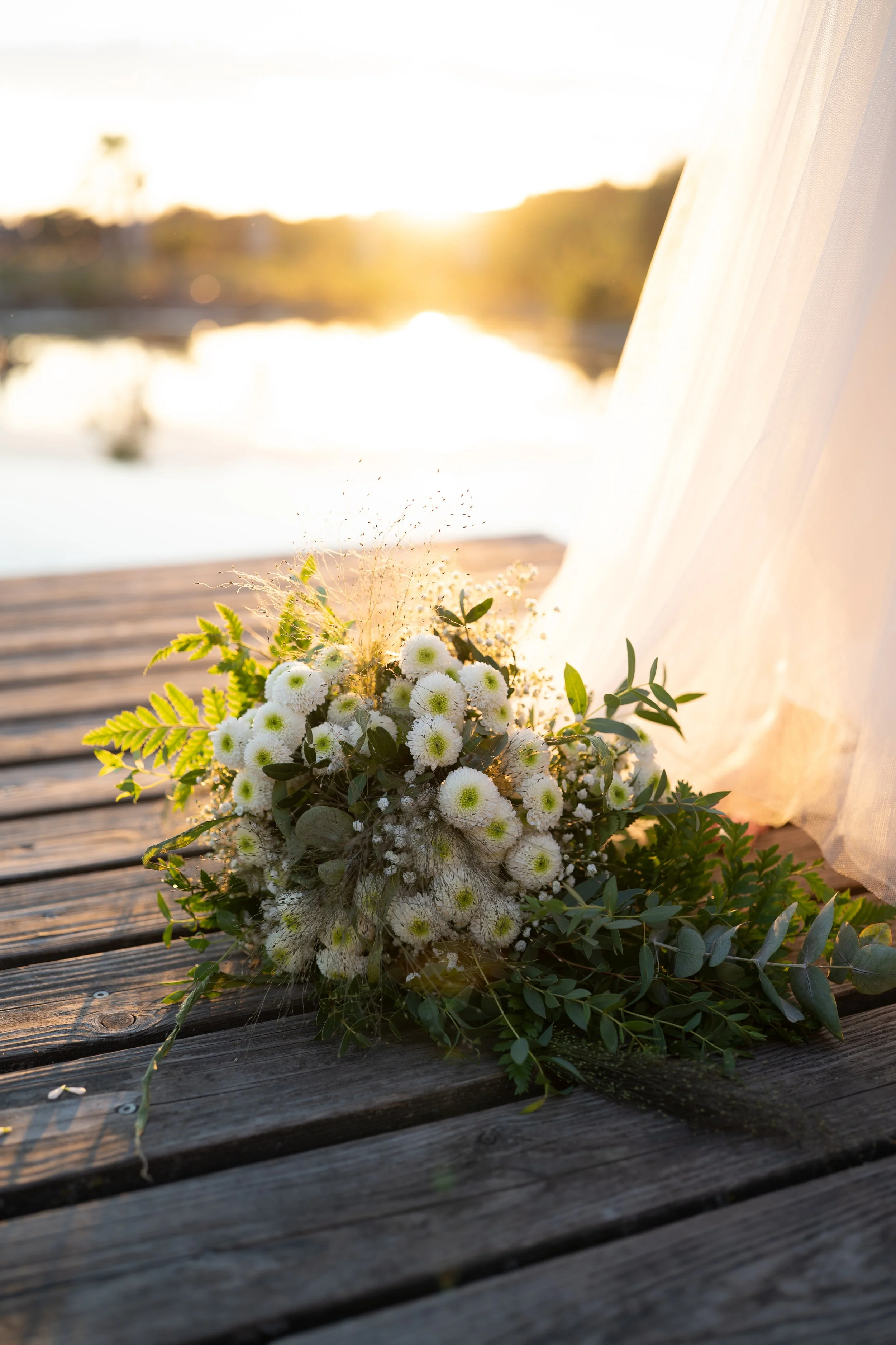 Ein Blumenstrauß mit weißen und grünen Blumen liegt auf einem Holzdeck, im Hintergrund die Sonne bei Sonnenuntergang über einem Wasser. Ein hölzerner Vorhang ist auf der rechten Seite sichtbar.