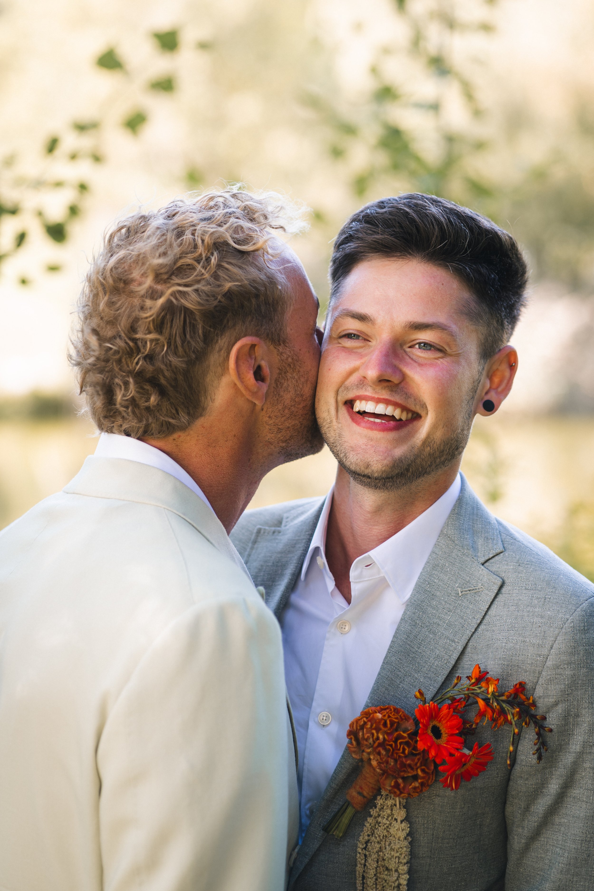 Zwei Männer bei einer Hochzeit, einer küsst den anderen auf die Wange, beide lächeln bei sonnigem Wetter im Freien.