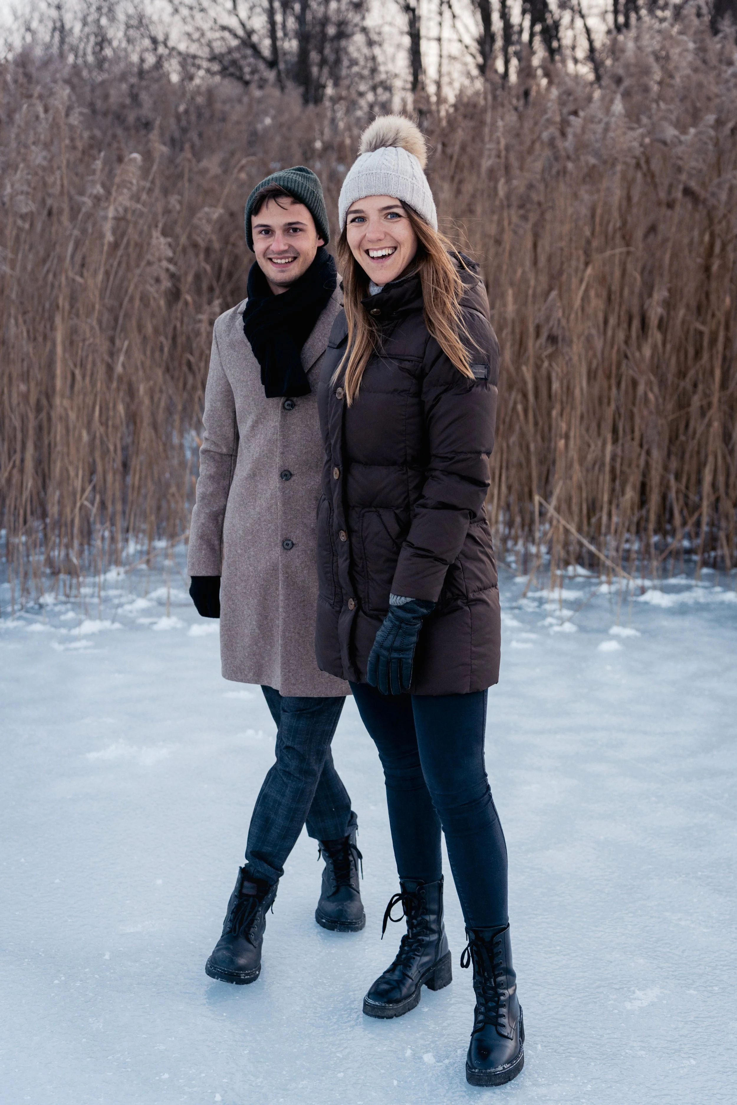 Zwei lachende Menschen, ein Mann und eine Frau, stehen auf einer Eisschicht im Winter, mit trockenem Schilf im Hintergrund. Sie tragen Winterkleidung, einschließlich Mäntel, Mützen und Handschuhe.