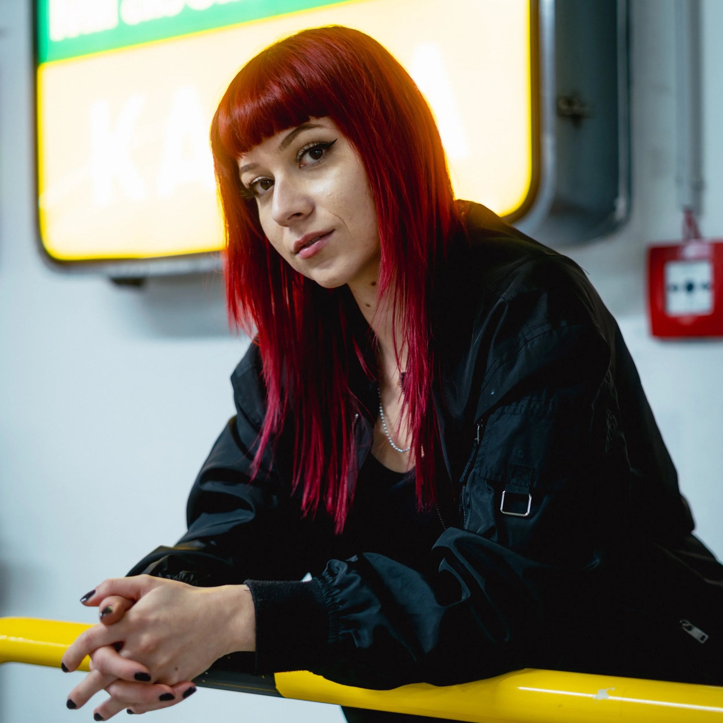 Junge Frau mit roten Haaren und schwarzer Jacke lehnt an einer gelben Haltestange in einem öffentlichen Verkehrsmittel, im Hintergrund ein gelbes Schild und ein rotes Feuerlöscher