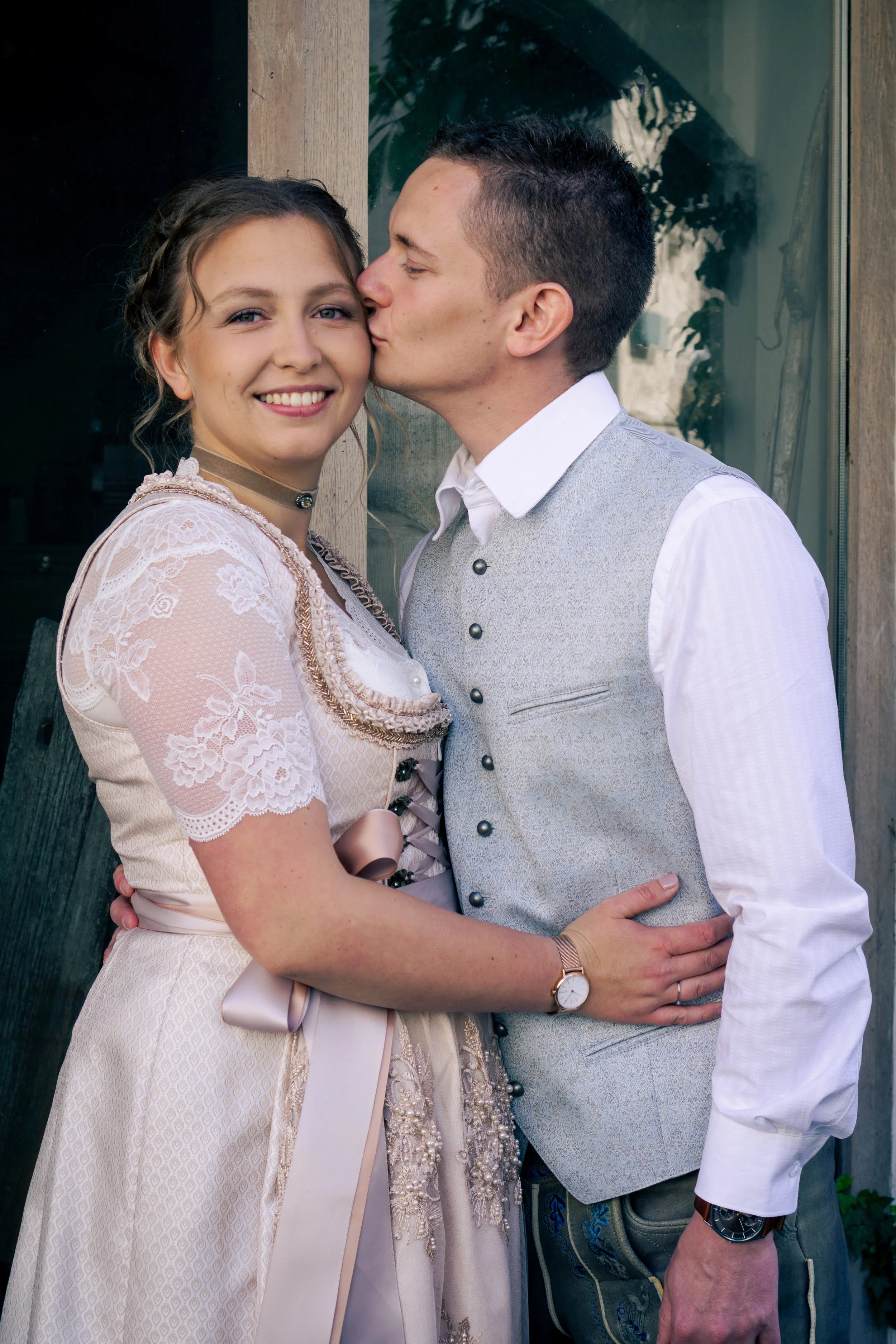 Ein junges Paar umarmt sich, der Mann küsst die Frau auf die Wange, beide tragen festliche Kleidung, im Hintergrund eine Fensterfront mit Pflanzen