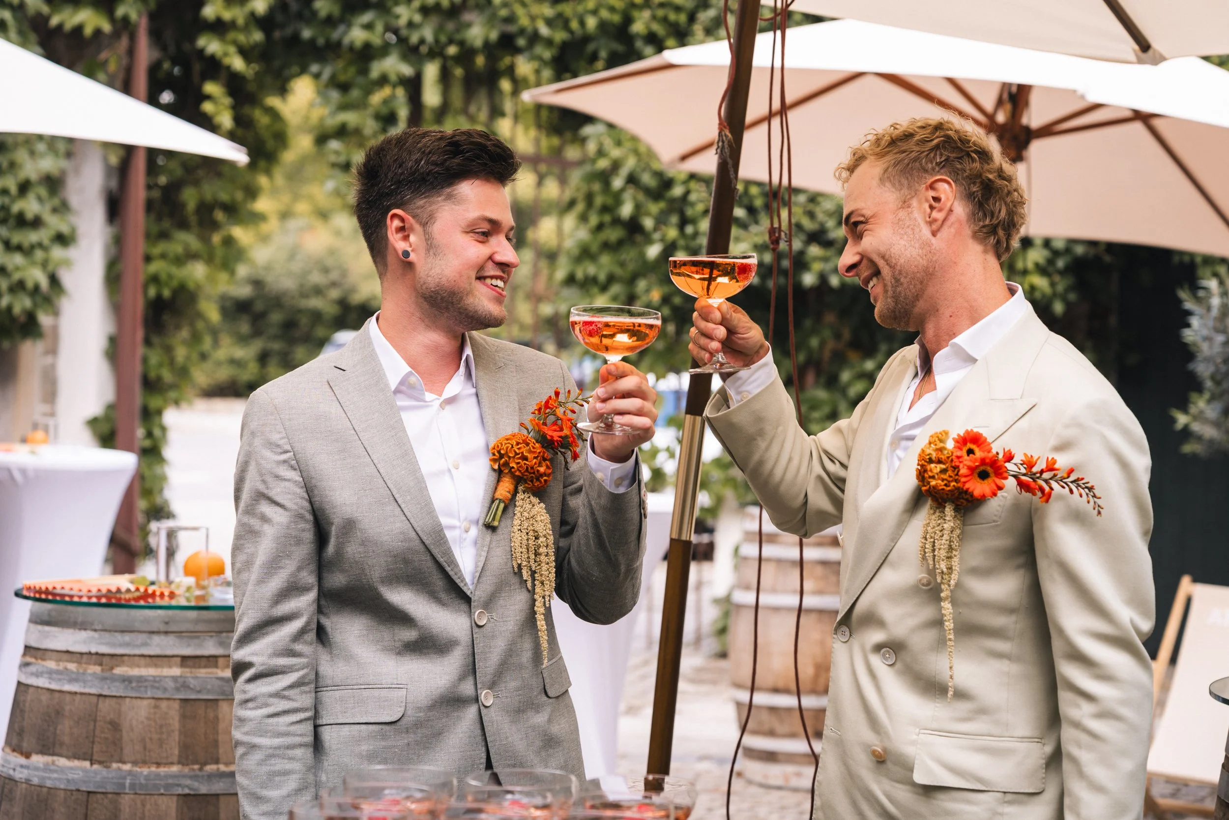 Zwei Männer in Anzügen mit Blumen auf der Brust stoßen bei einer Feier mit rosafarbigen Getränken an.