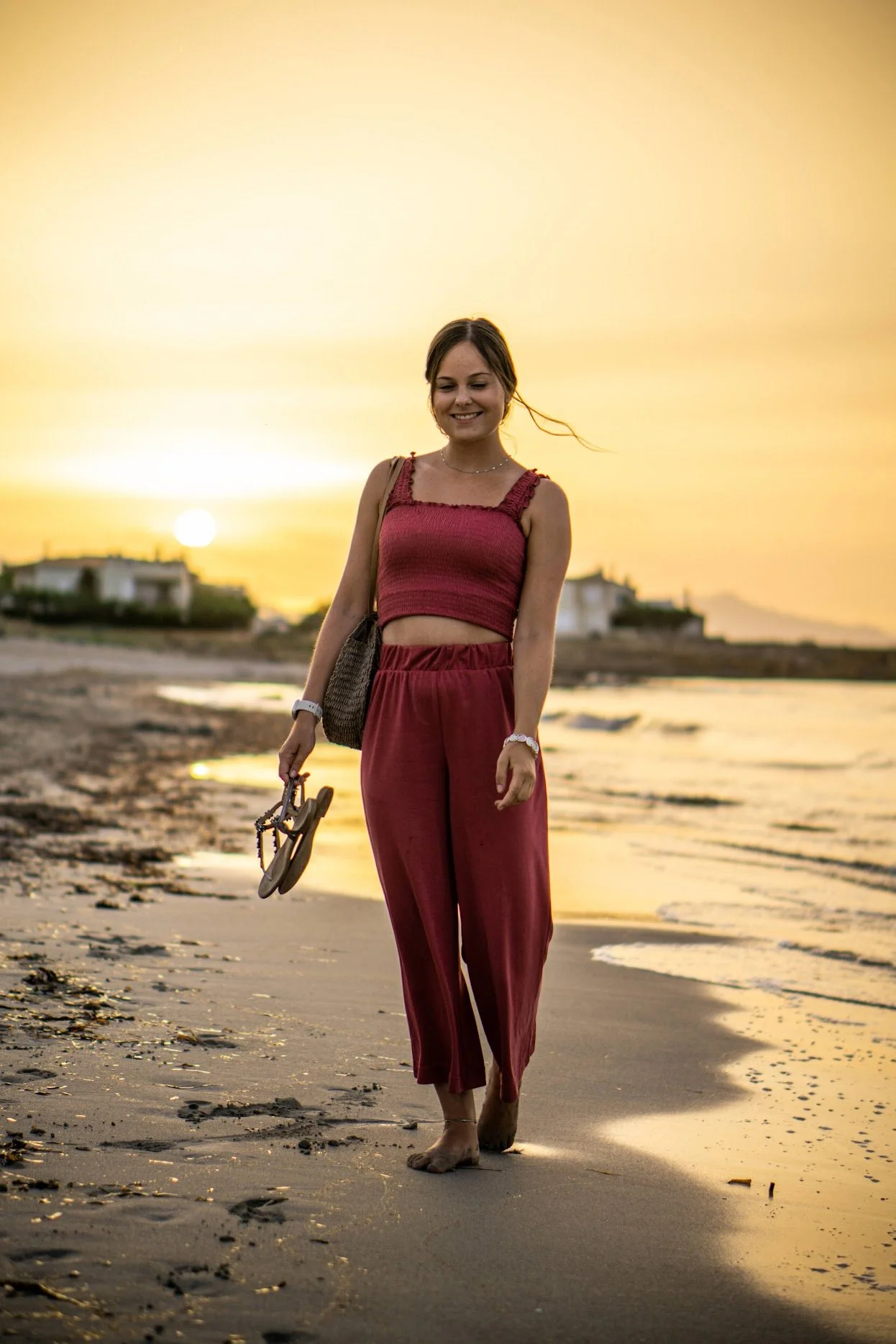 Eine lachende junge Frau in einem roten Top und roten Hosen, die barfuß am Strand entlangspaziert, bei Sonnenuntergang, mit Sonnenbrille in der Hand.
