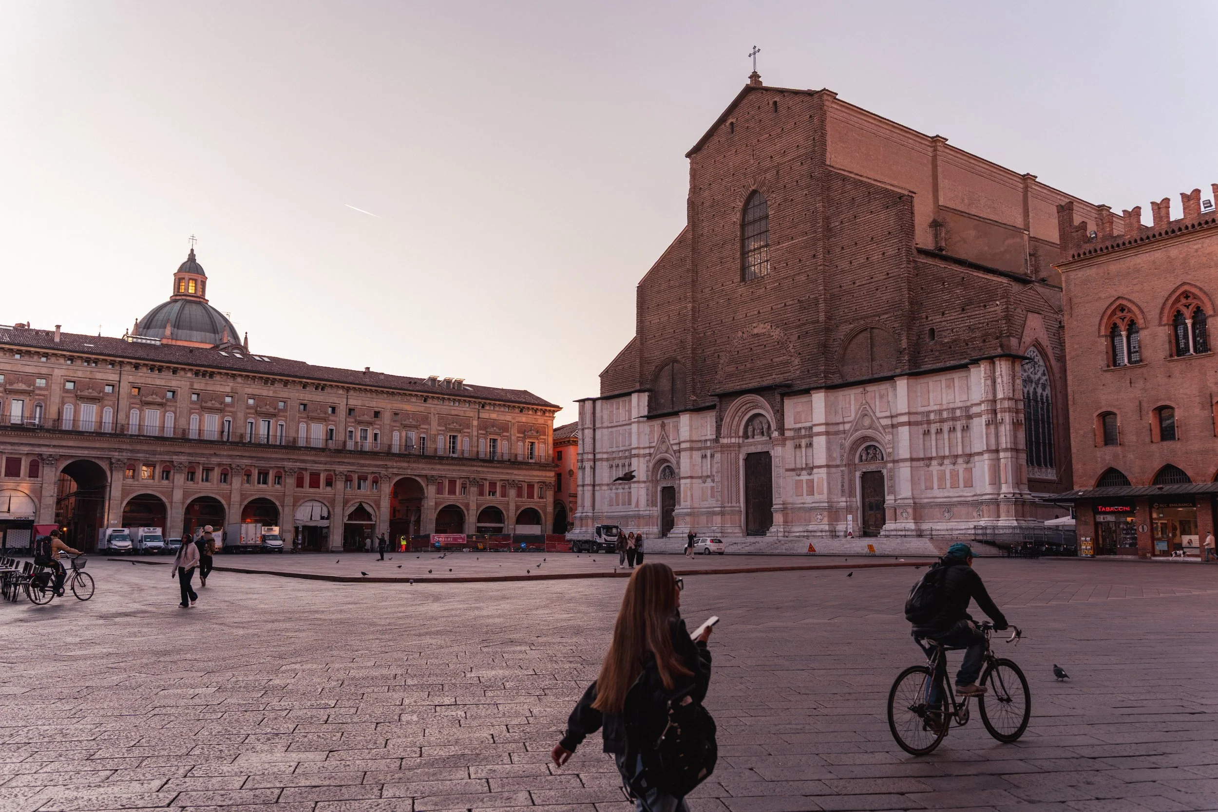 A photo of a historic brick cathedral with a marble facade, situated in a city square with people walking and cycling during sunset.