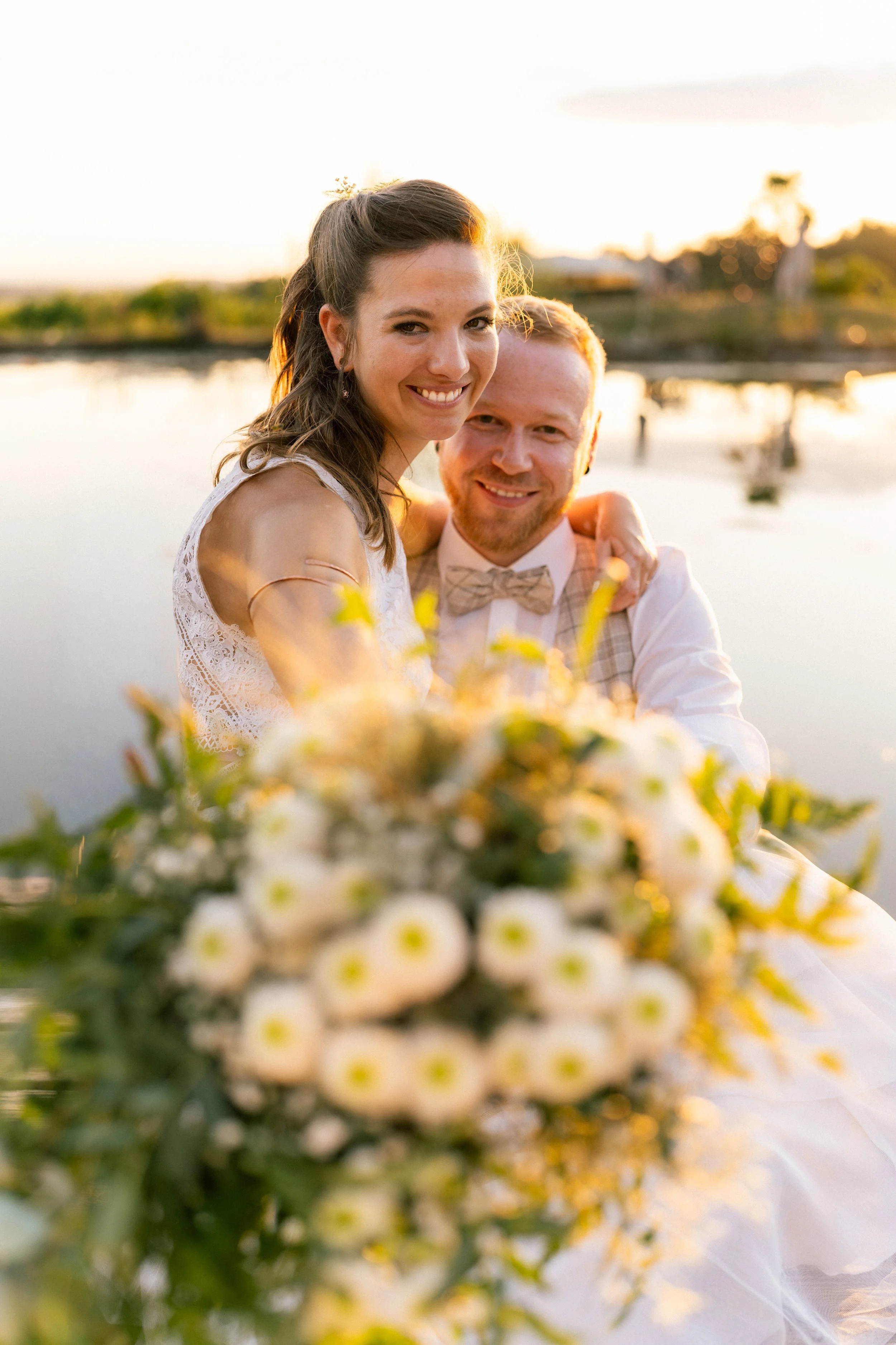 Ein glückliches Paar während einer Hochzeit im Freien bei Sonnenuntergang in Salzburg, mit Wasser im Hintergrund und bunten Blumen im Vordergrund.
