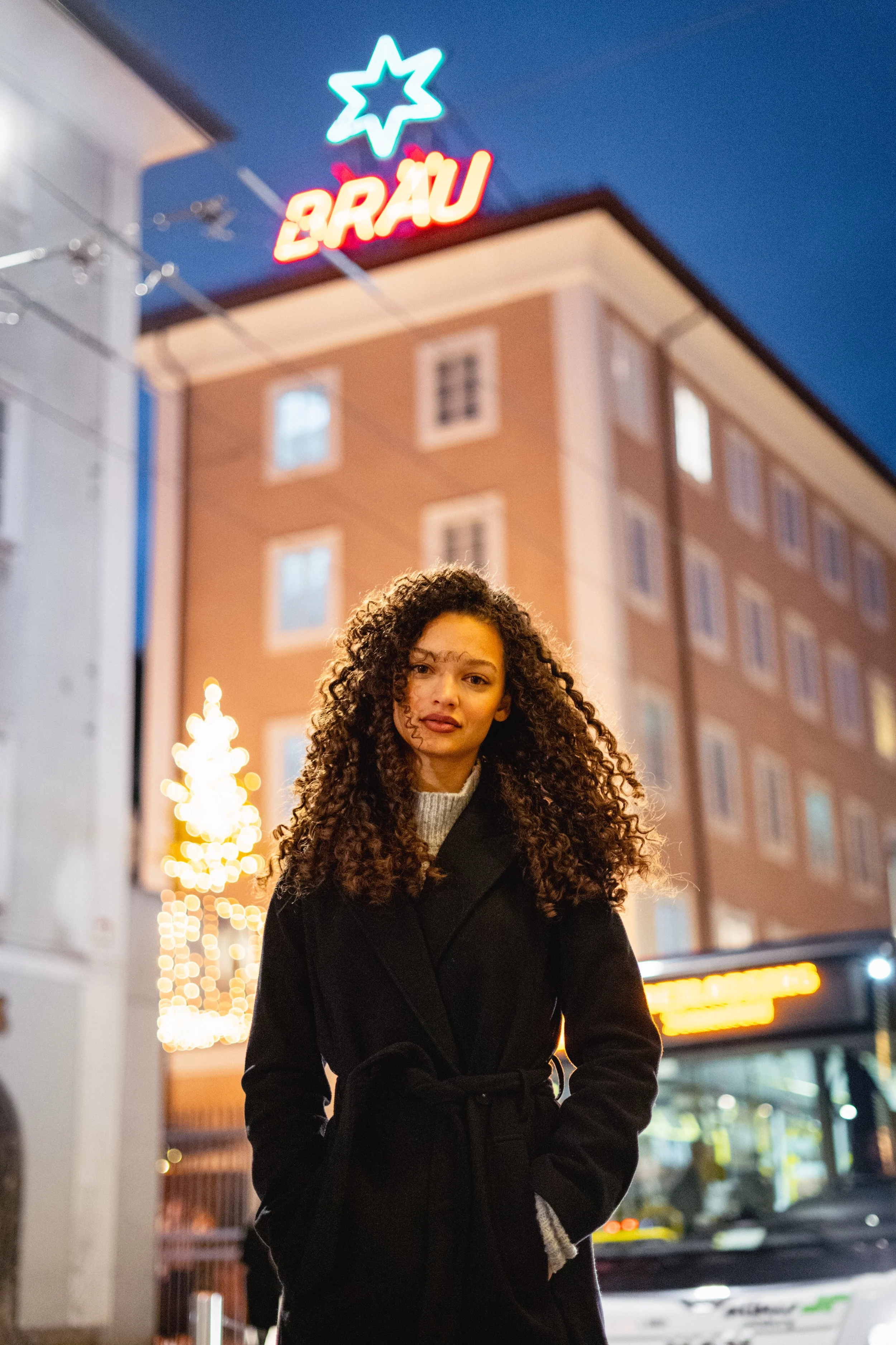 Eine Frau mit lockigem Haar trägt einen schwarzen Mantel auf einer belebten Straße bei Nacht, im Hintergrund sind bunte Lichter und ein Gebäude mit einem Neonzeichen mit der Aufschrift 'BRÄU' sichtbar.