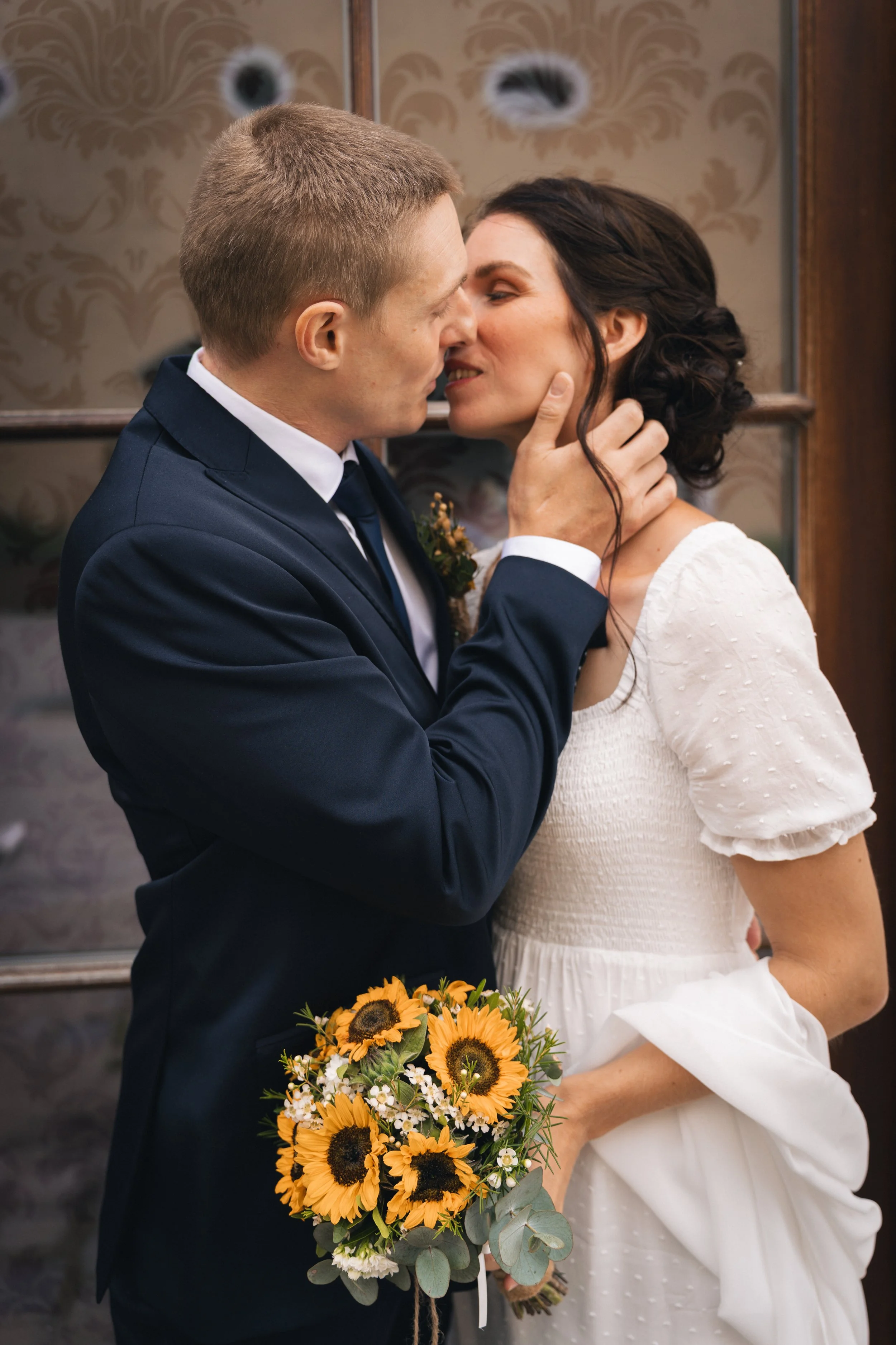 Ein Brautpaar in formeller Kleidung, die sich küsst, während die Braut einen Sonnenblumenstrauß hält.