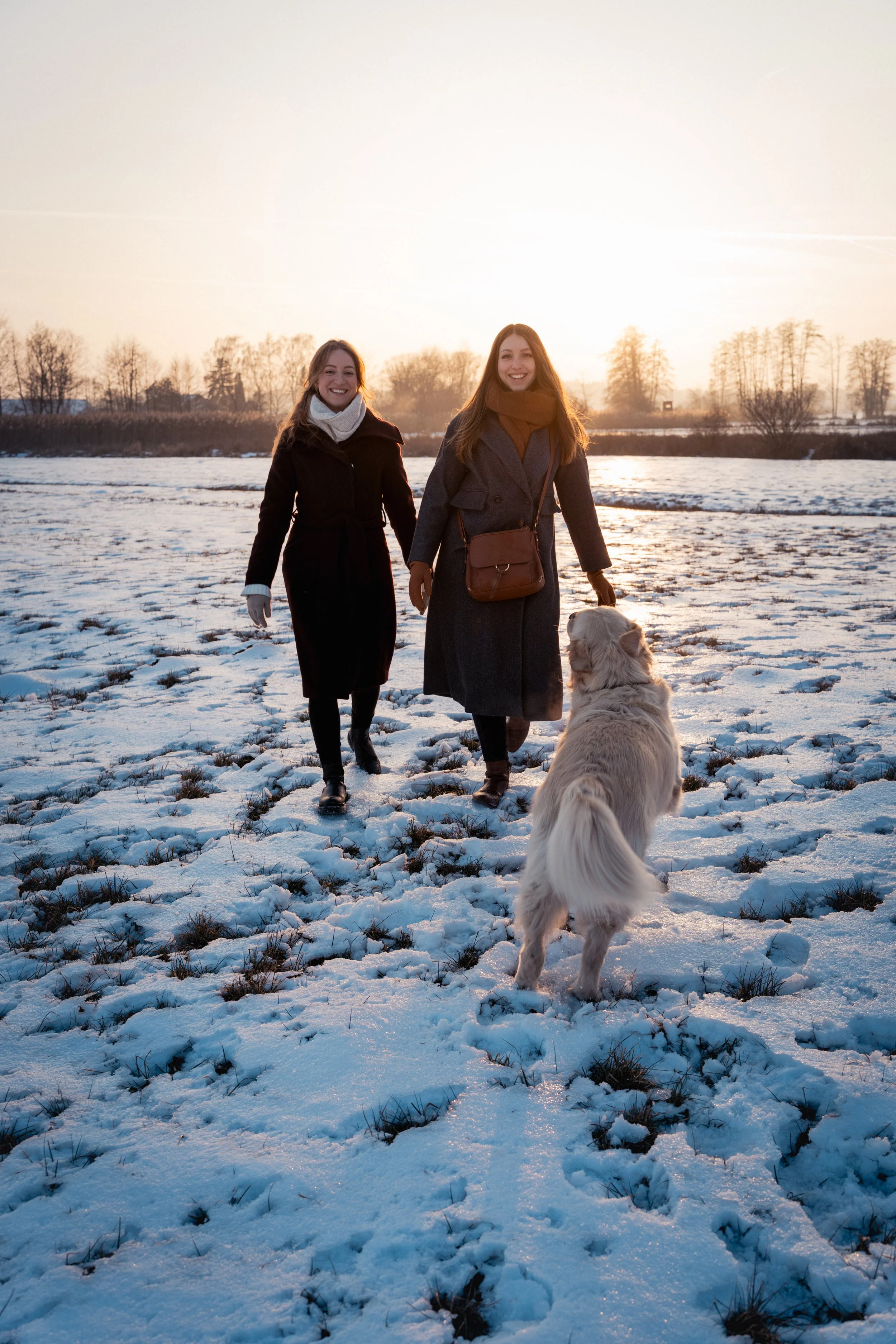 Zwei lachende Frauen in Winterkleidung gehen im Schnee spazieren, während ein Hund auf sie zukommt, bei Sonnenuntergang im Hintergrund.