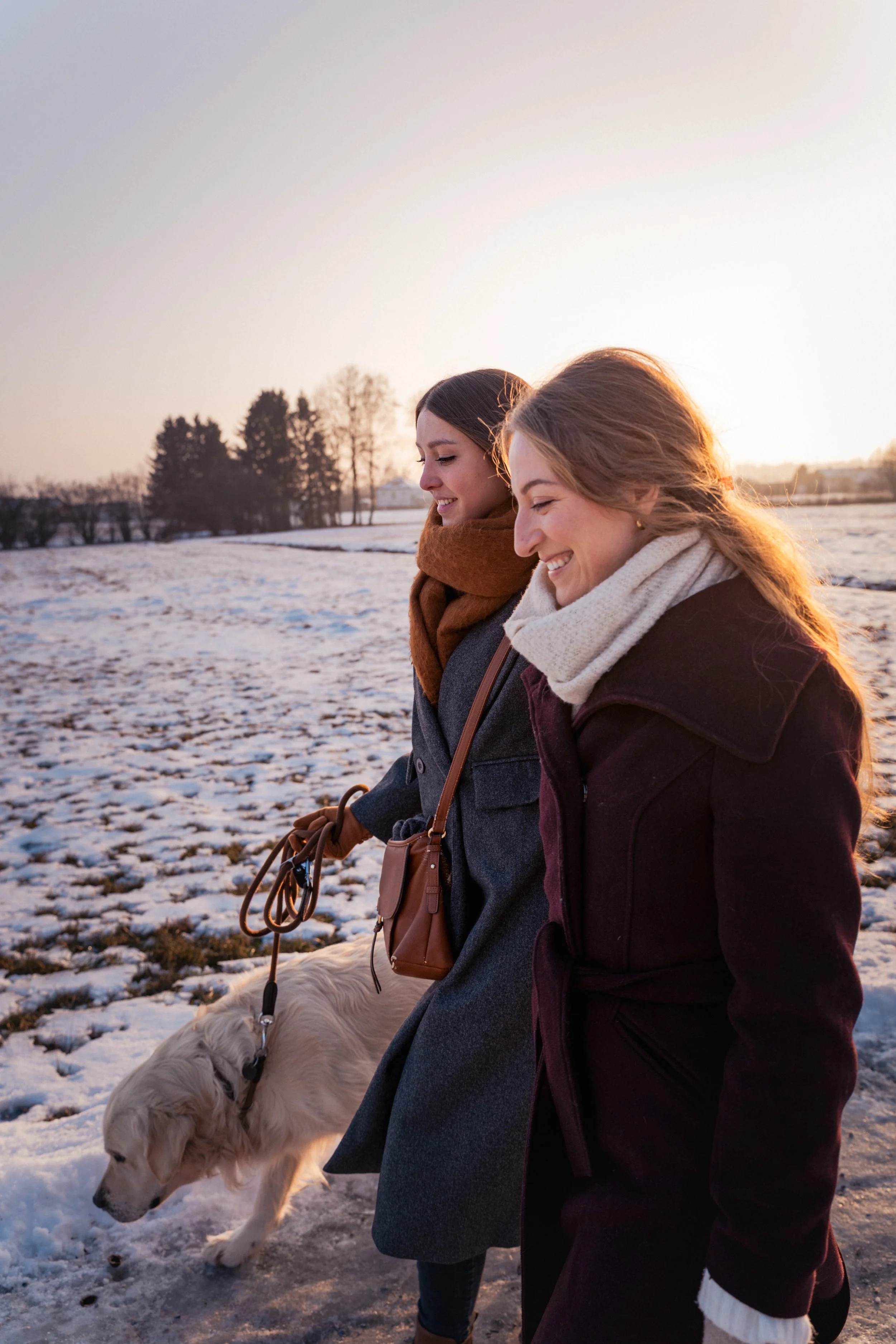 Zwei lachende Frauen gehen im Winter spazieren, begleitet von einem Hund, bei Sonnenuntergang, in einer verschneiten Landschaft.