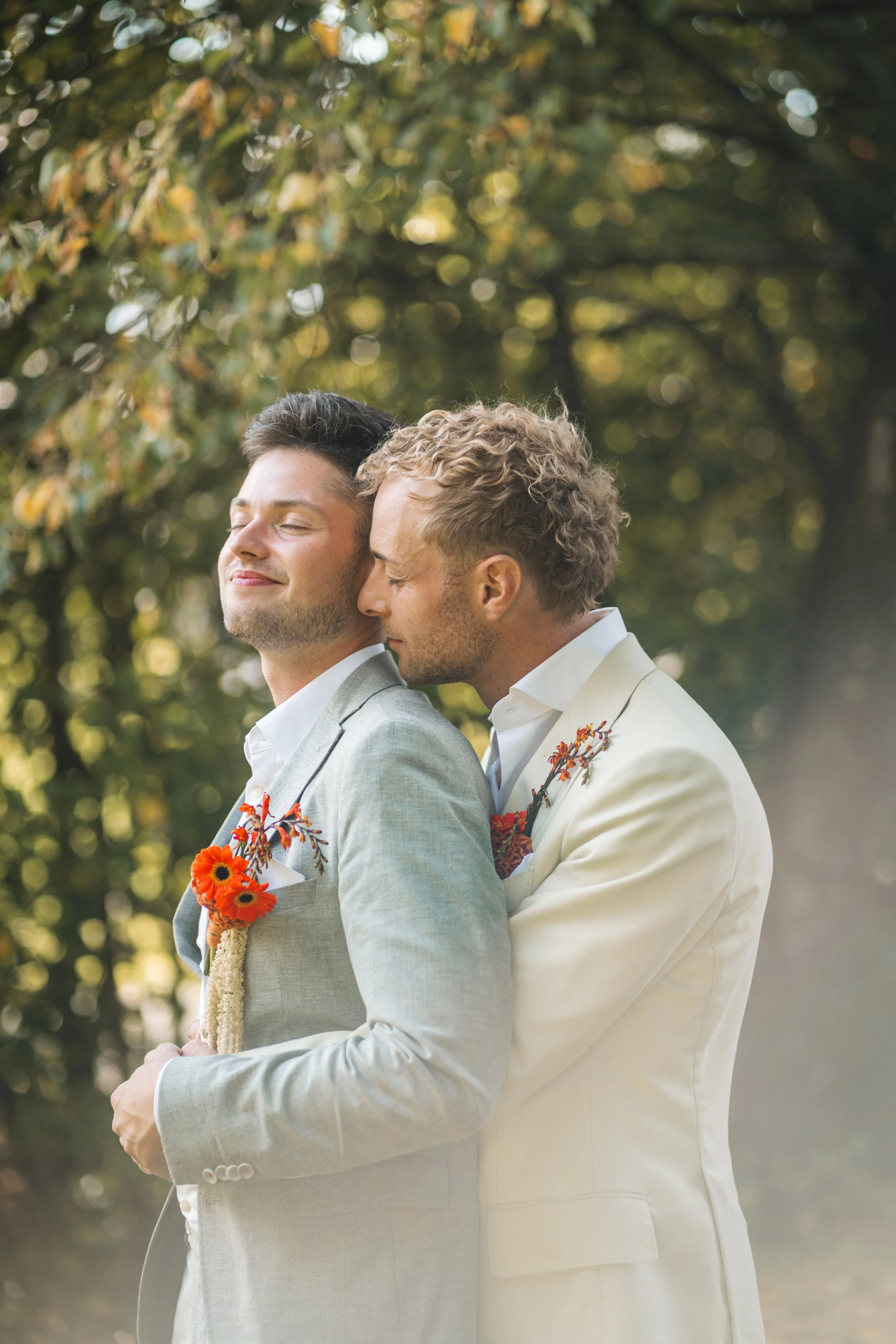 Zwei Männer in Anzügen, die bei einer Hochzeitszeremonie im Freien stehen, einer umarmt den anderen von hinten, alle beide lächelnd, mit fröhlicher Atmosphäre im Hintergrund.