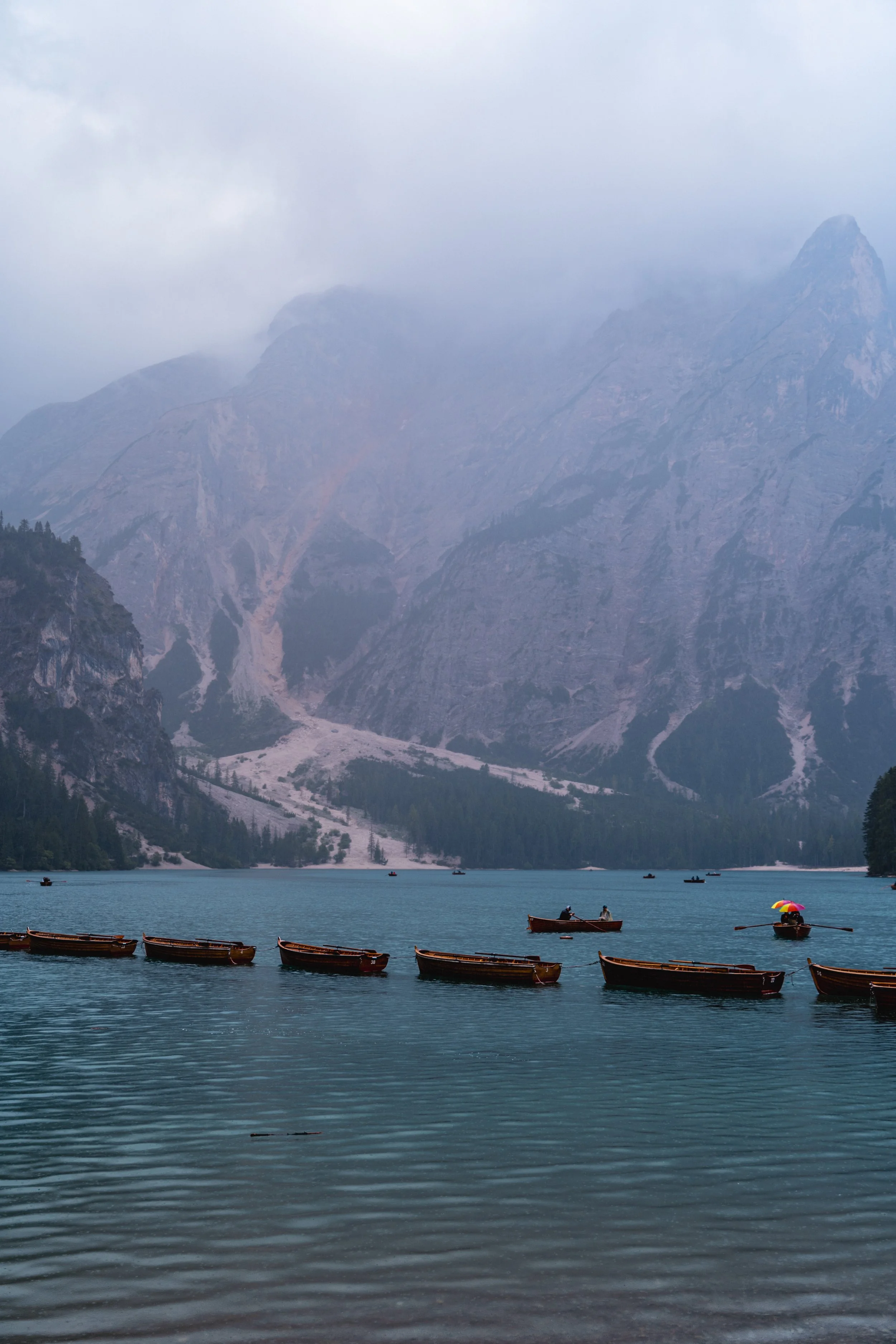 Blick auf einen wolkenverhangenen Bergsee mit Booten im Wasser, umgeben von Bergen und dichten Wäldern