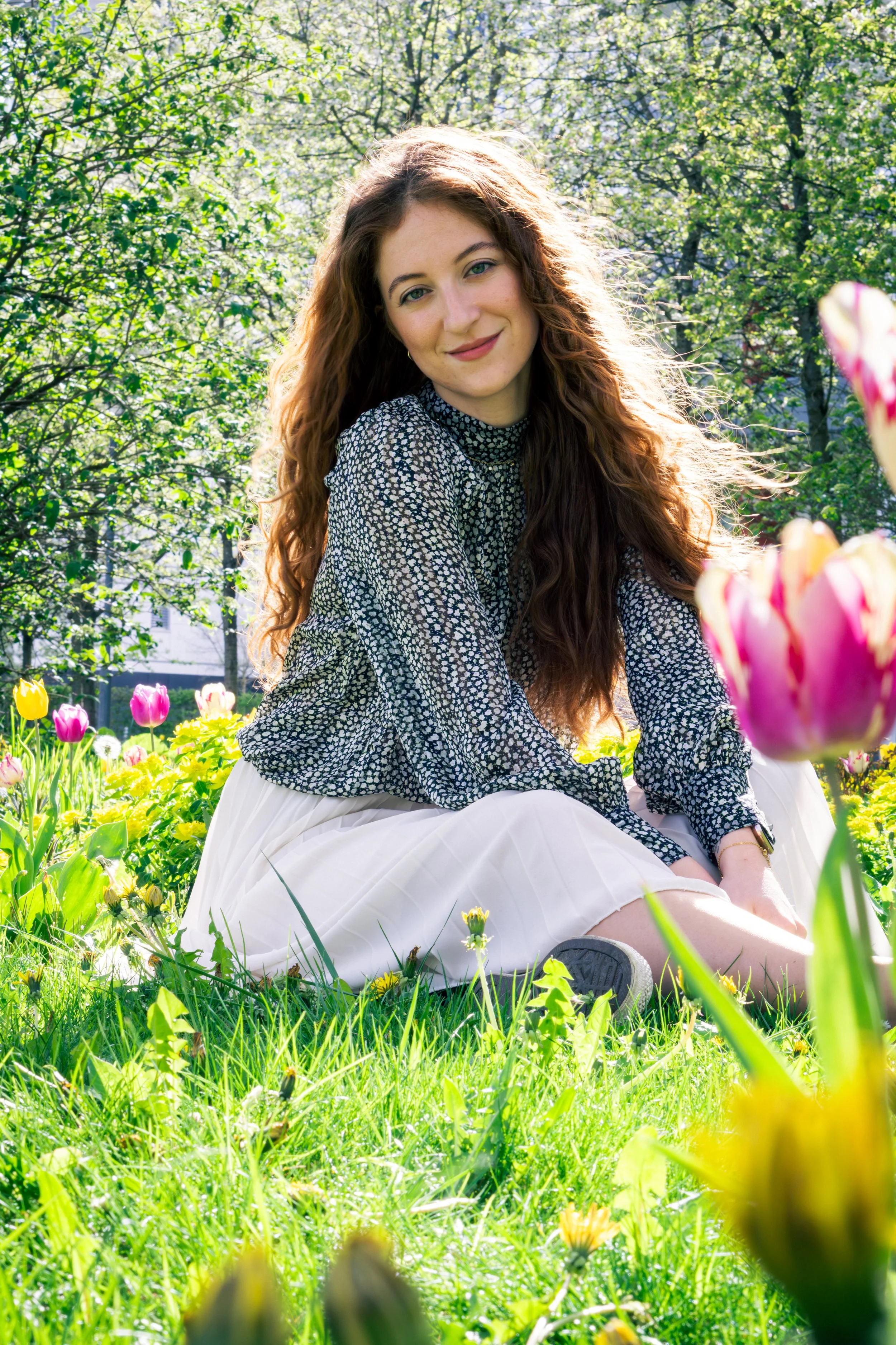 Junge Frau sitzt im Garten mit bunten Tulpen und grünen Pflanzen im Hintergrund, lacht und genießt die Sonnenstrahlen.