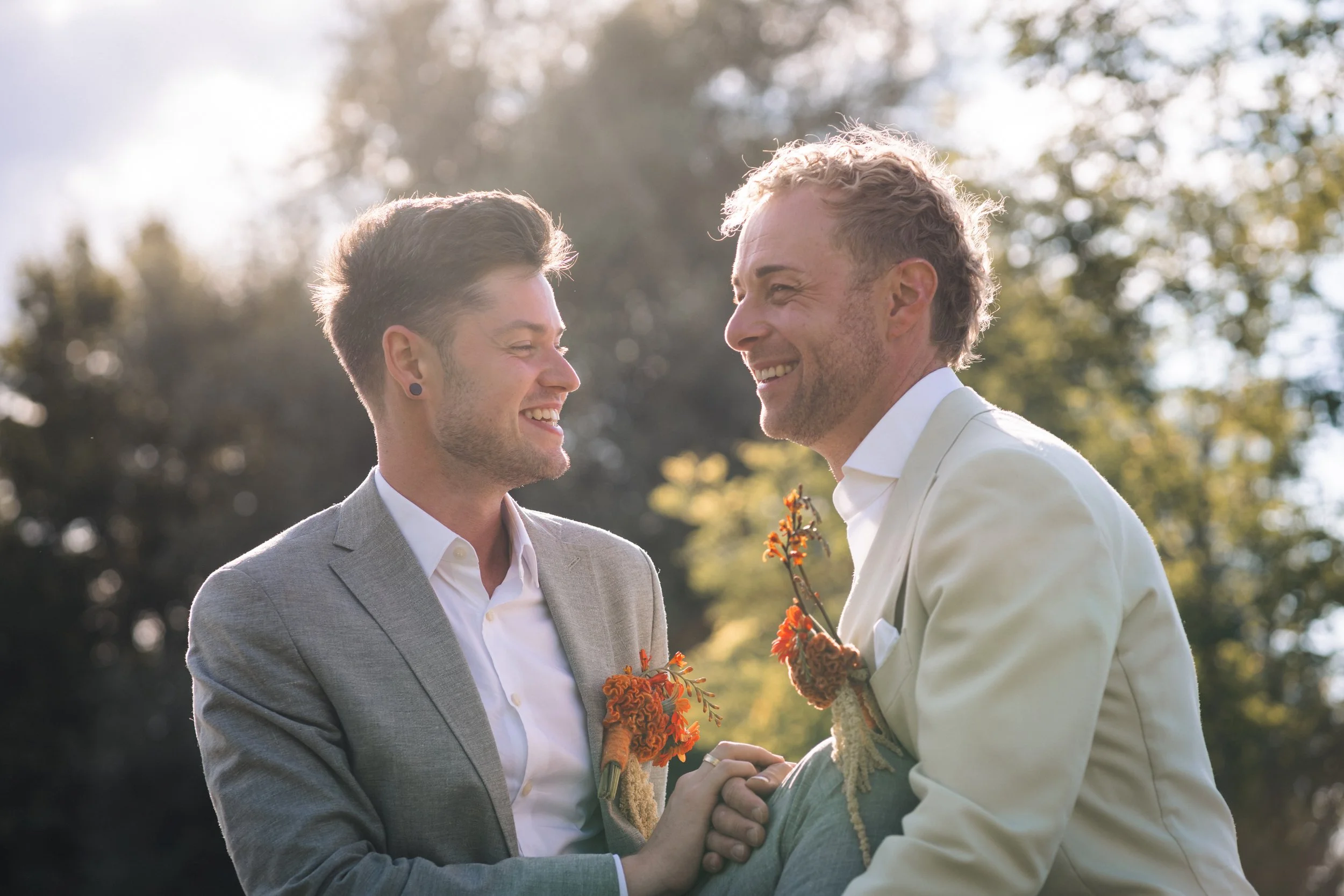 Zwei Männer, die sich bei einer Hochzeit im Freien anlächelnd die Hände halten. Einer trägt einen grauen Anzug, der andere einen weißen Anzug, beide mit Blumen im Revers. Im Hintergrund sind Bäume und Sonnenlicht sichtbar.