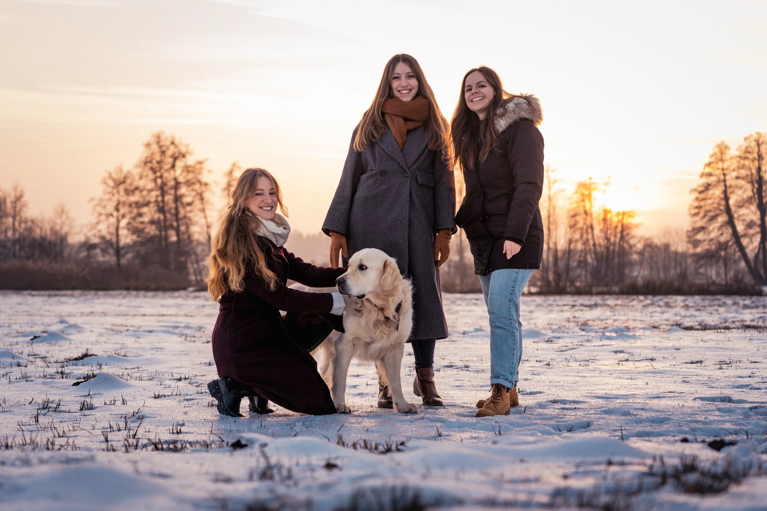 Drei junge Frauen mit einem Golden Retriever im Schnee bei Sonnenuntergang im Freien.