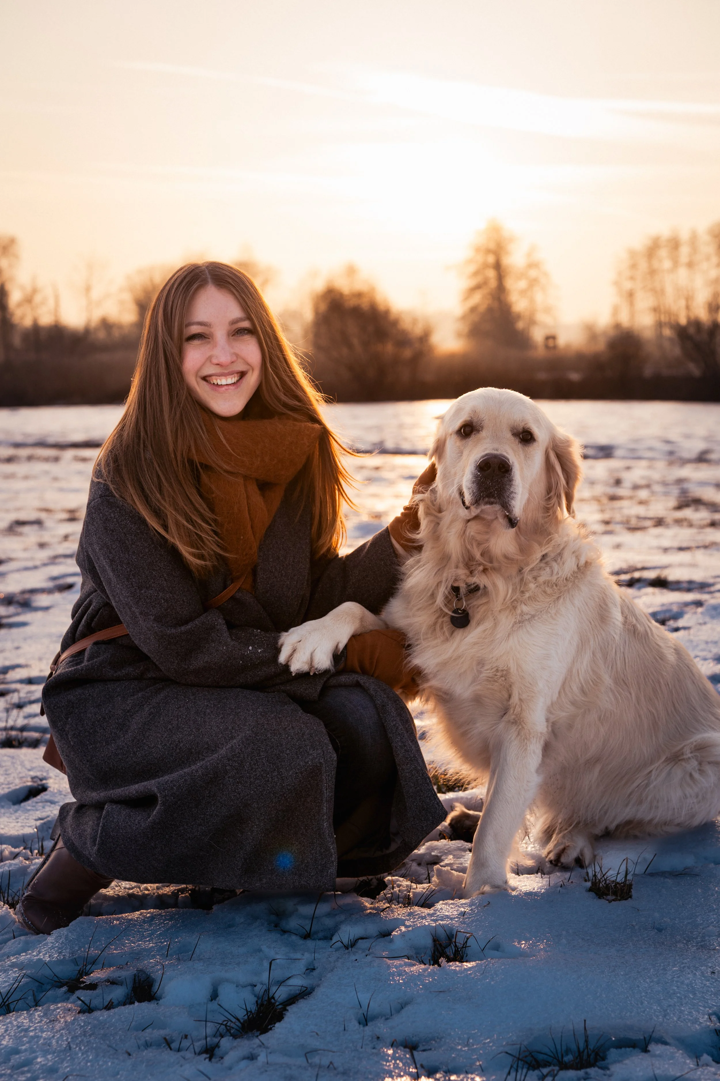 Eine lachende Frau mit braunem Schal neben einem Golden Retriever im Schnee bei Sonnenuntergang.