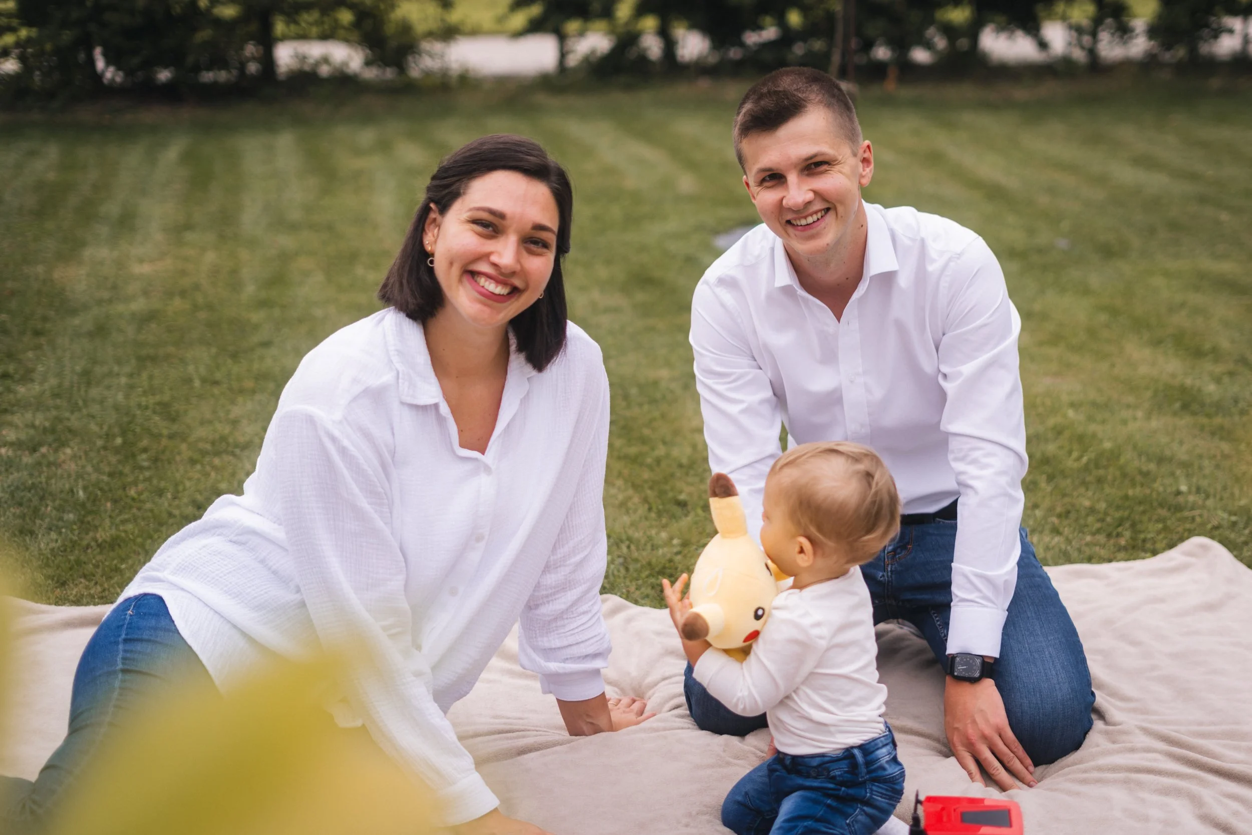 Familie mit zwei Erwachsenen und einem kleinen Kind sitzt auf einer Decke im Park, das Kind spielt mit einer Plüschpferd. Es ist sonnig, und die Familie sieht glücklich aus.