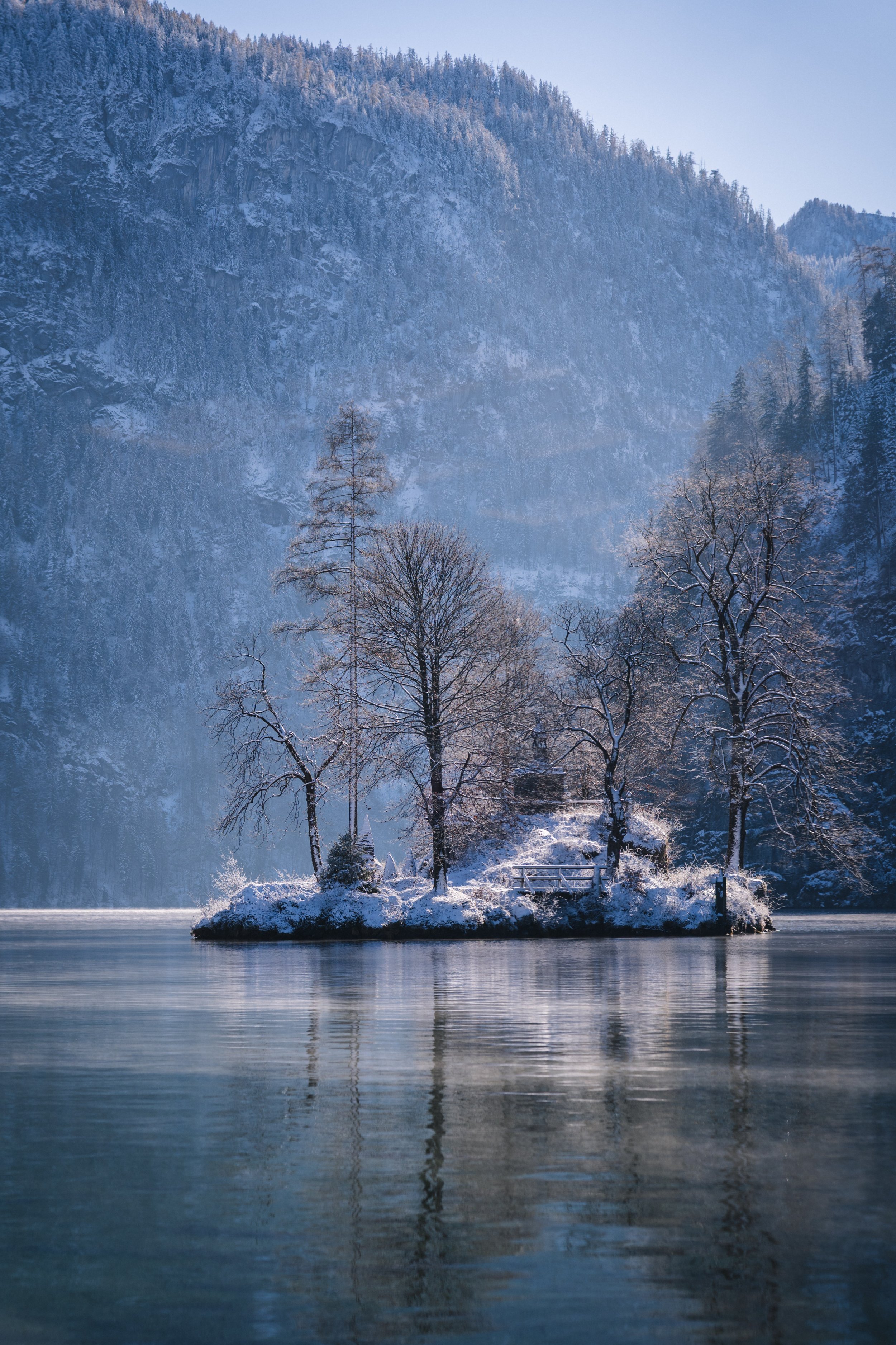 Ein kleiner schneebedeckter Insel mit Bäumen und einem kleinen Bauwerk, umgeben von einem ruhigen See mit Bergen im Hintergrund im winterlichen Szenario.