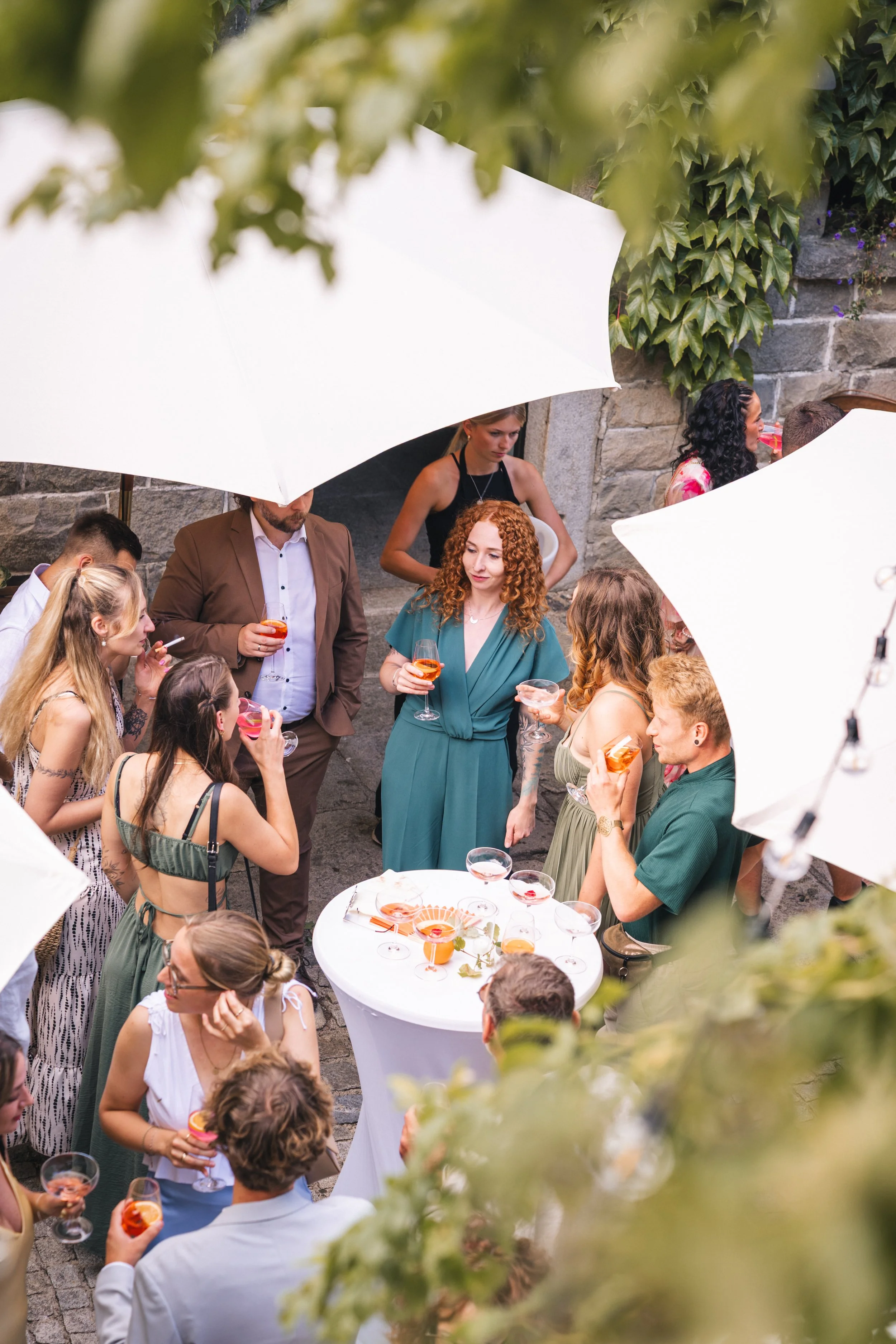 Menschen bei einer Gartenparty, die Cocktails trinken, im Freien, umgeben von Pflanzen und weißen Sonnenschirmen.