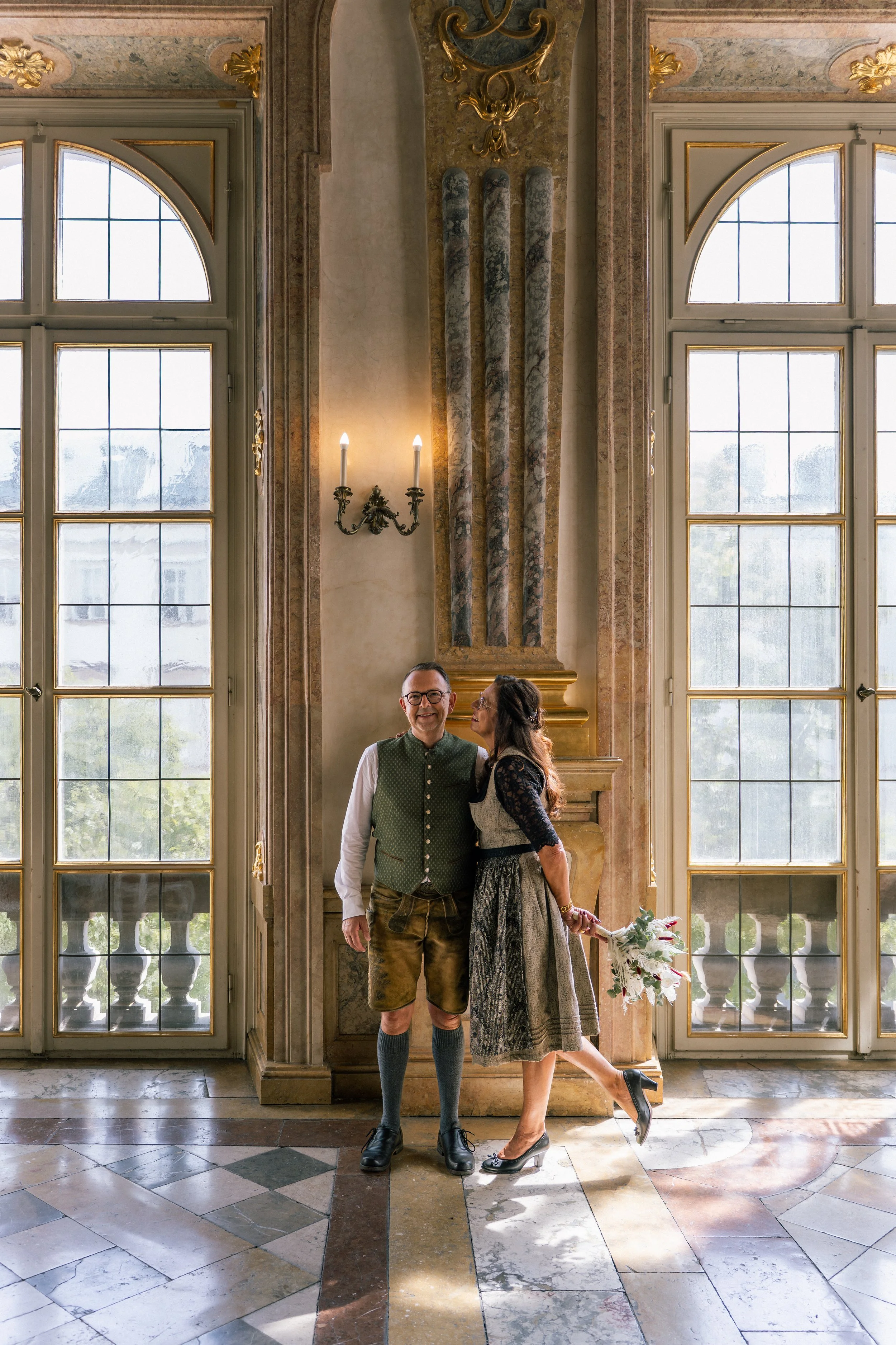 Eine Braut und ein Bräutigam stehen in einem eleganten Saal mit großen Fenstern, einem Marmorboden und antiker Dekoration im Trauungssaal im Schloss Mirabell in Salzburg.