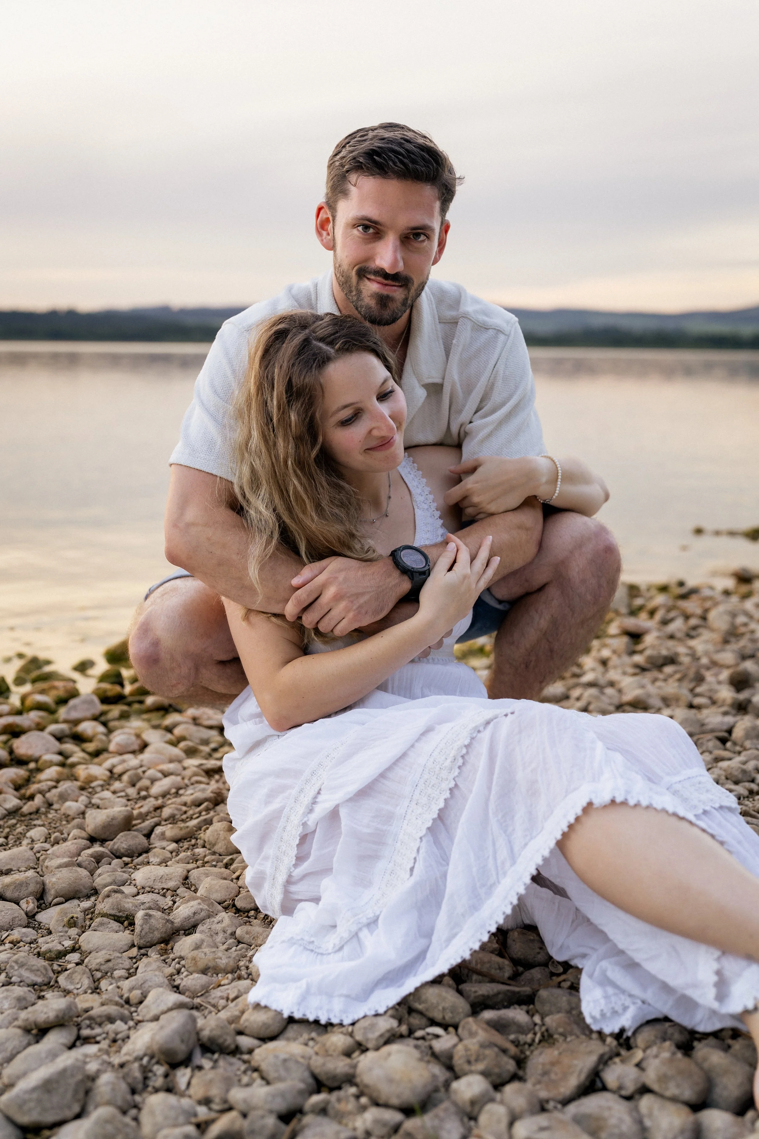 Ein Paar, vermutlich ein Mann und eine Frau, sitzen am Strand mit Kieselsteinen, umarmt und entspannt, während die Sonne untergeht oder scheint, mit einem ruhigen Wasserhintergrund und Hügel in der Ferne.