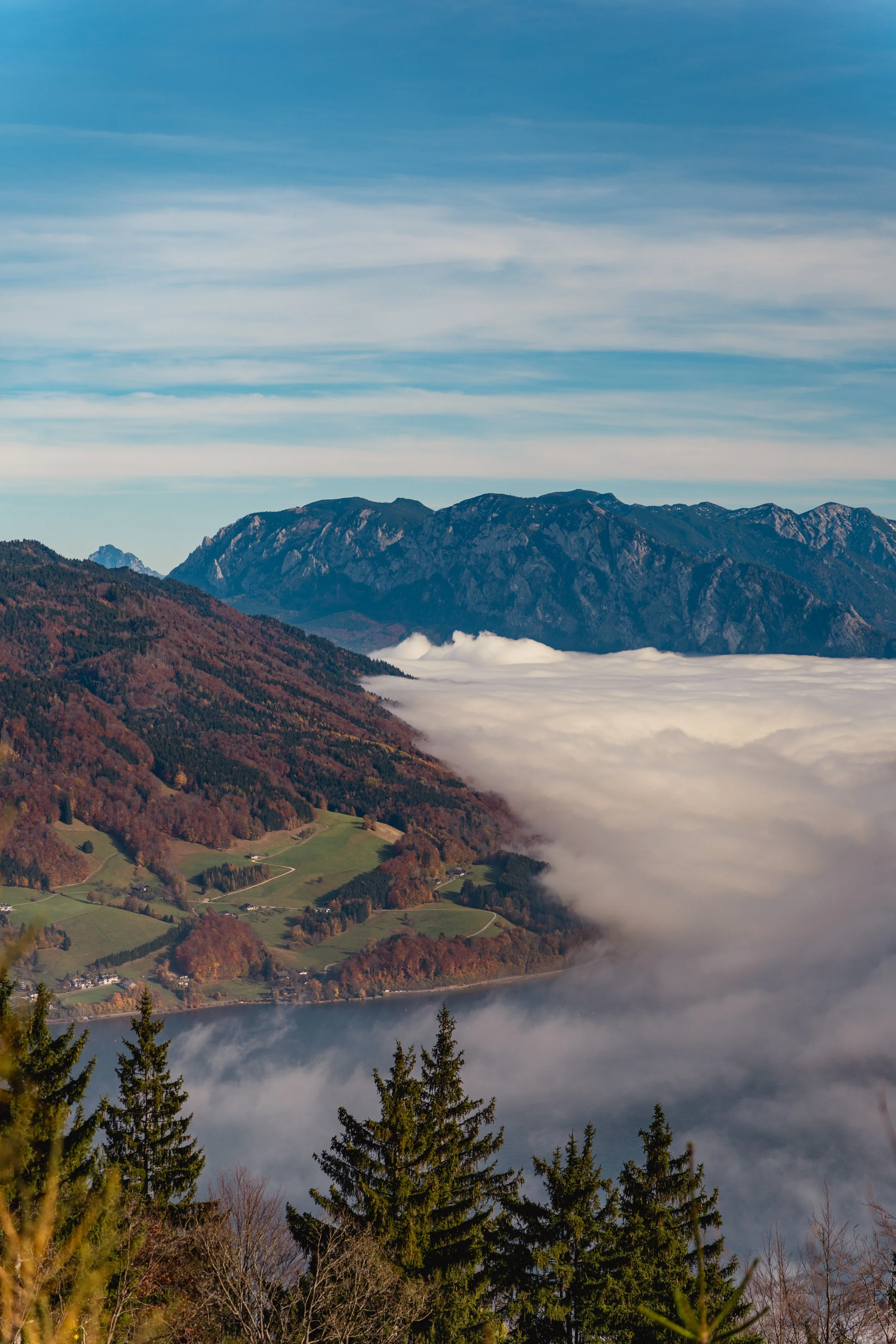 Berglandschaft mit Wolken im Tal, grün bewaldete Hügel und im Hintergrund hohe Berge unter einem blauen Himmel.