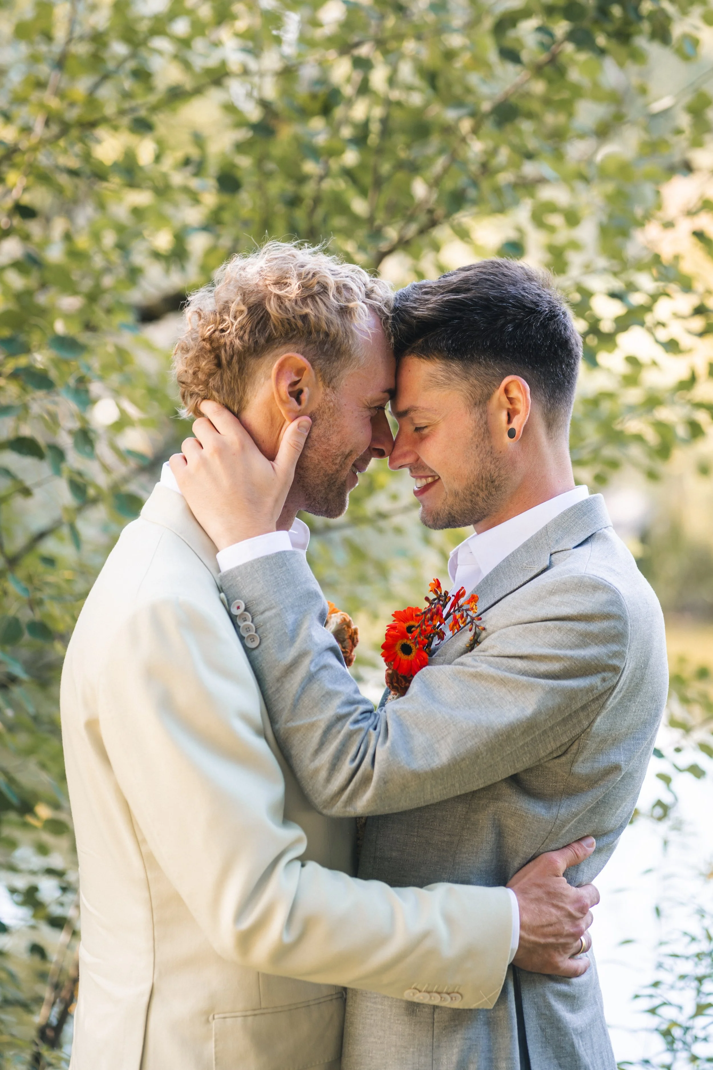 Zwei Männer in Anzügen, die sich liebevoll berühren und ihre Stirn aneinanderlegen, in einer natürlichen Umgebung mit grünen Blättern im Hintergrund.