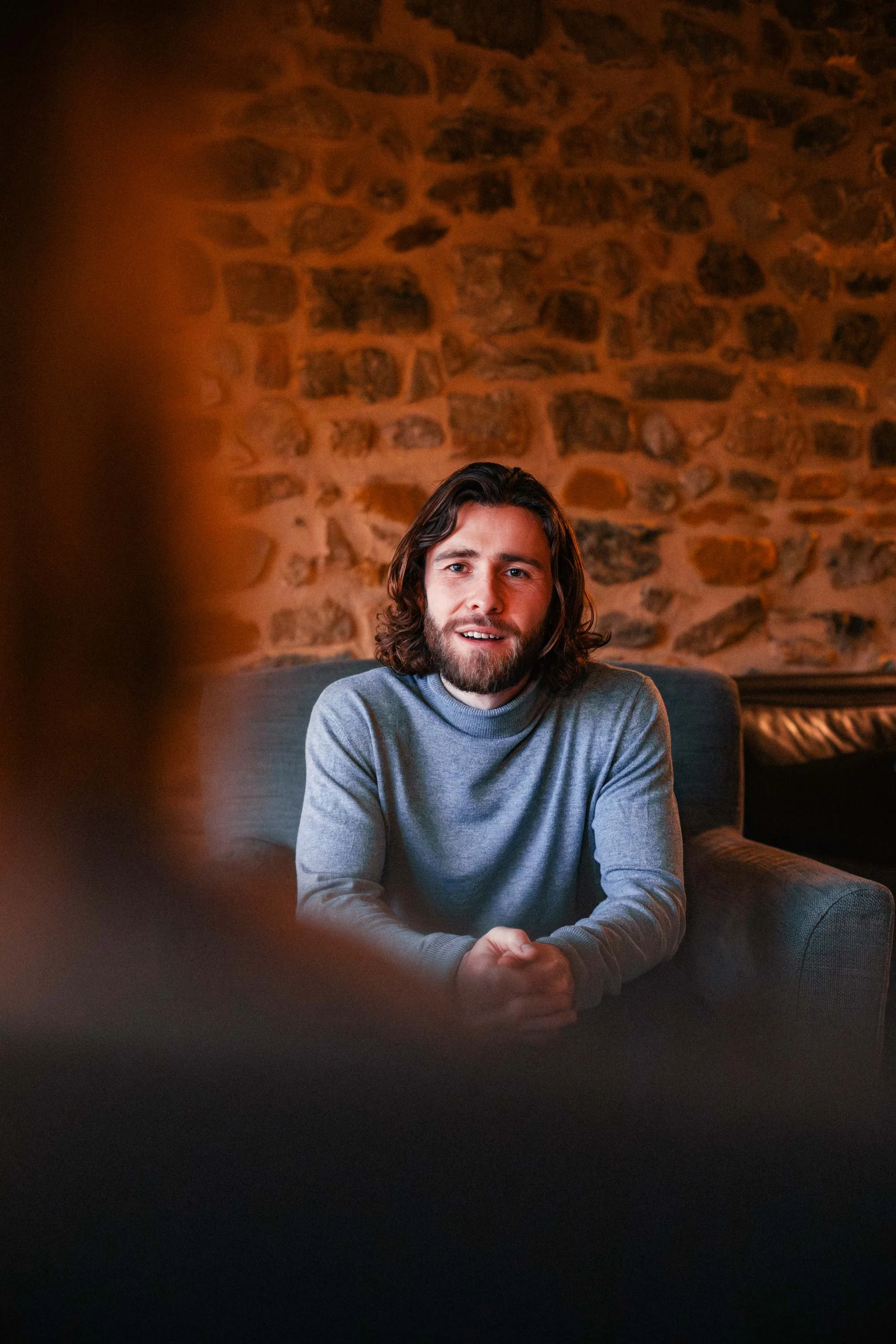 Un homme avec des cheveux longs et une barbe, assis sur un canapé en cuir, dans une pièce avec un mur en pierre en arrière-plan, parle ou écoute, dans une ambiance chaleureuse.