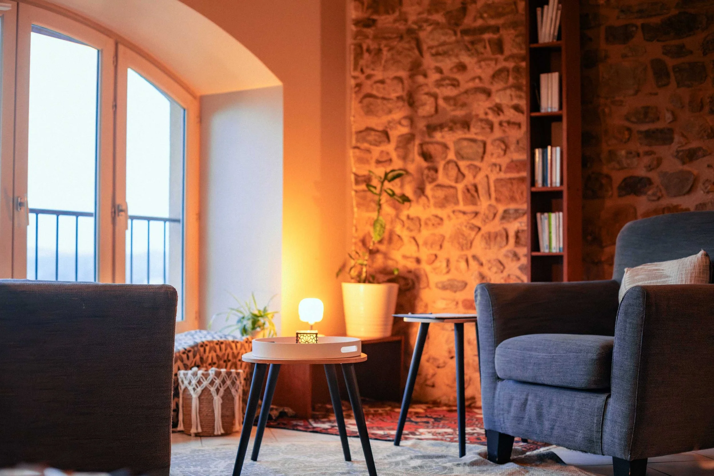 Salon cosy avec fauteuils gris, table basse, mur en Pierre, étagère avec livres, plantes, lampes et fenêtre avec balcon.