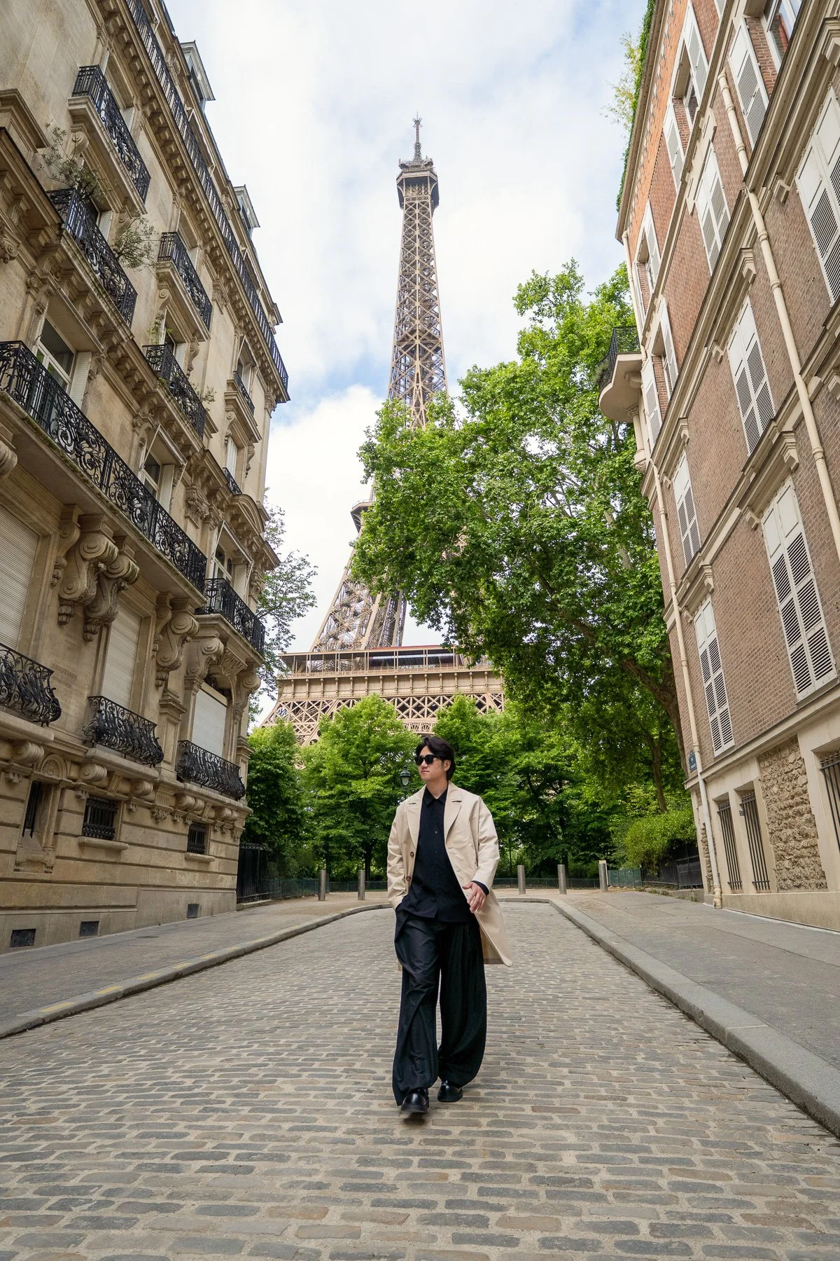 A person with black hair, sunglasses, a beige trench coat, black shirt, and black wide-leg pants walking on a cobblestone street with trees and buildings, the Eiffel Tower is visible in the background.