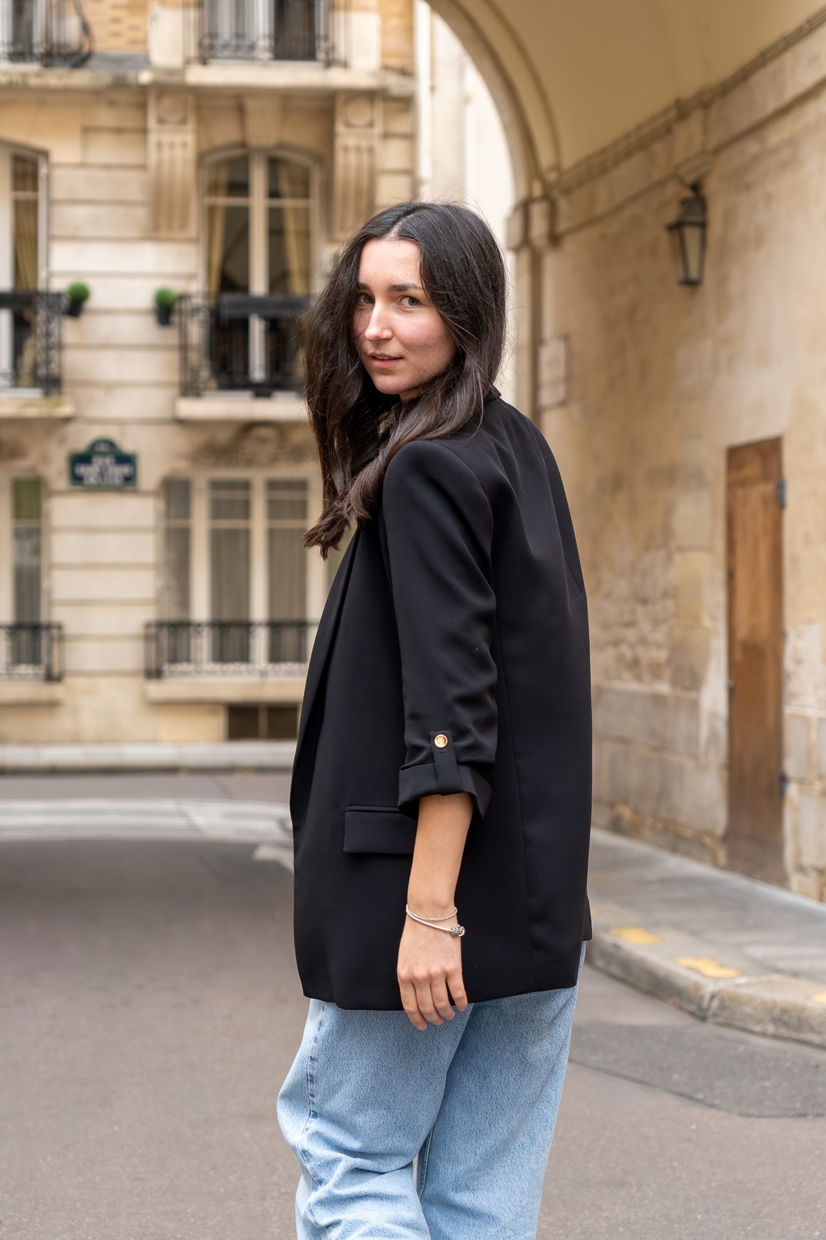 A woman with dark hair in a black blazer and light blue jeans standing on a city street, with European-style buildings and balconies in the background.