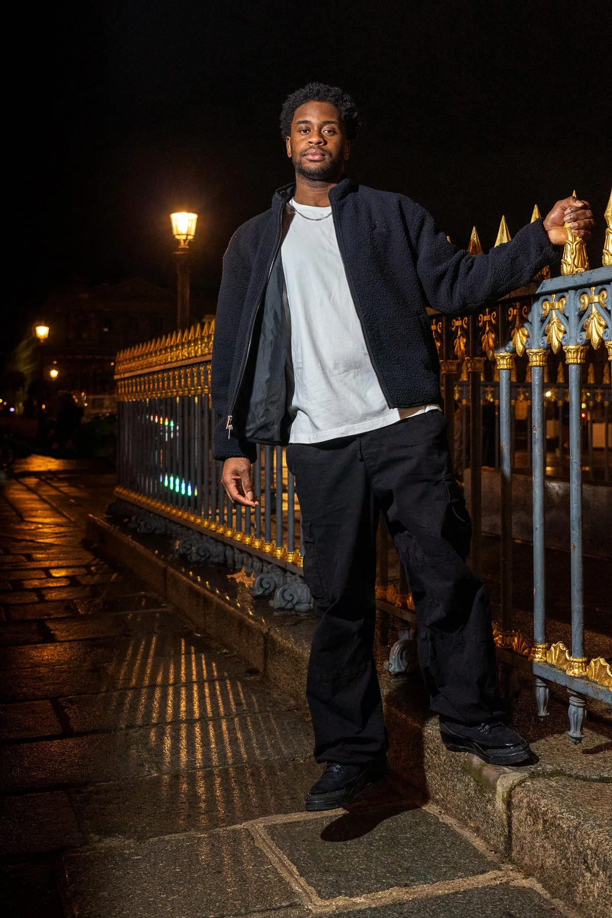 A young man stands at night on a street with a decorative iron fence, illuminated by street lamps, wearing a black jacket, white t-shirt, black cargo pants, and black shoes.