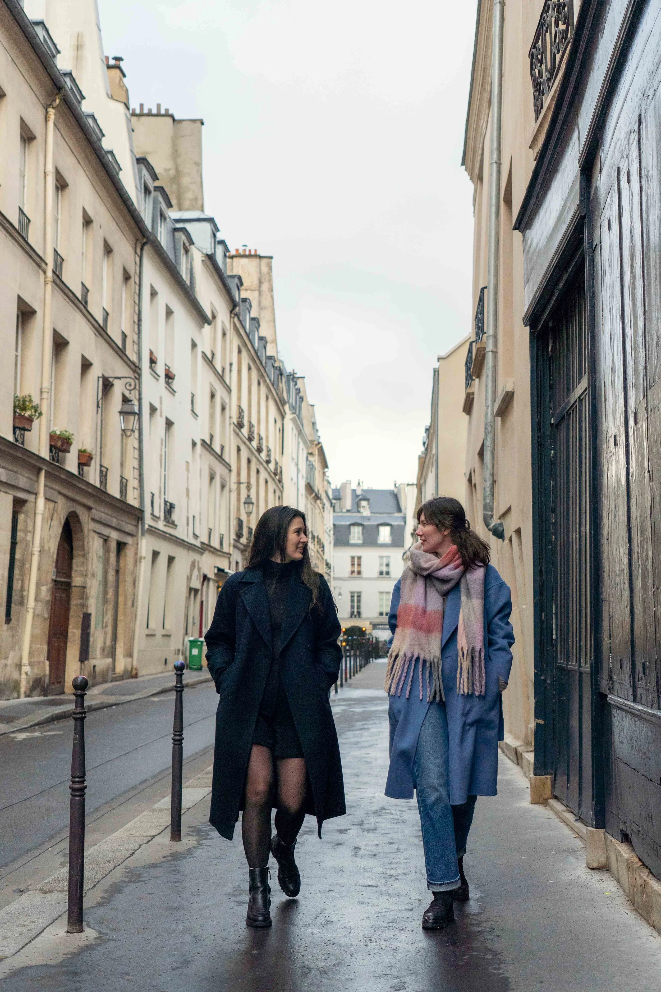 Two women walking and talking on a city sidewalk, surrounded by European-style buildings.