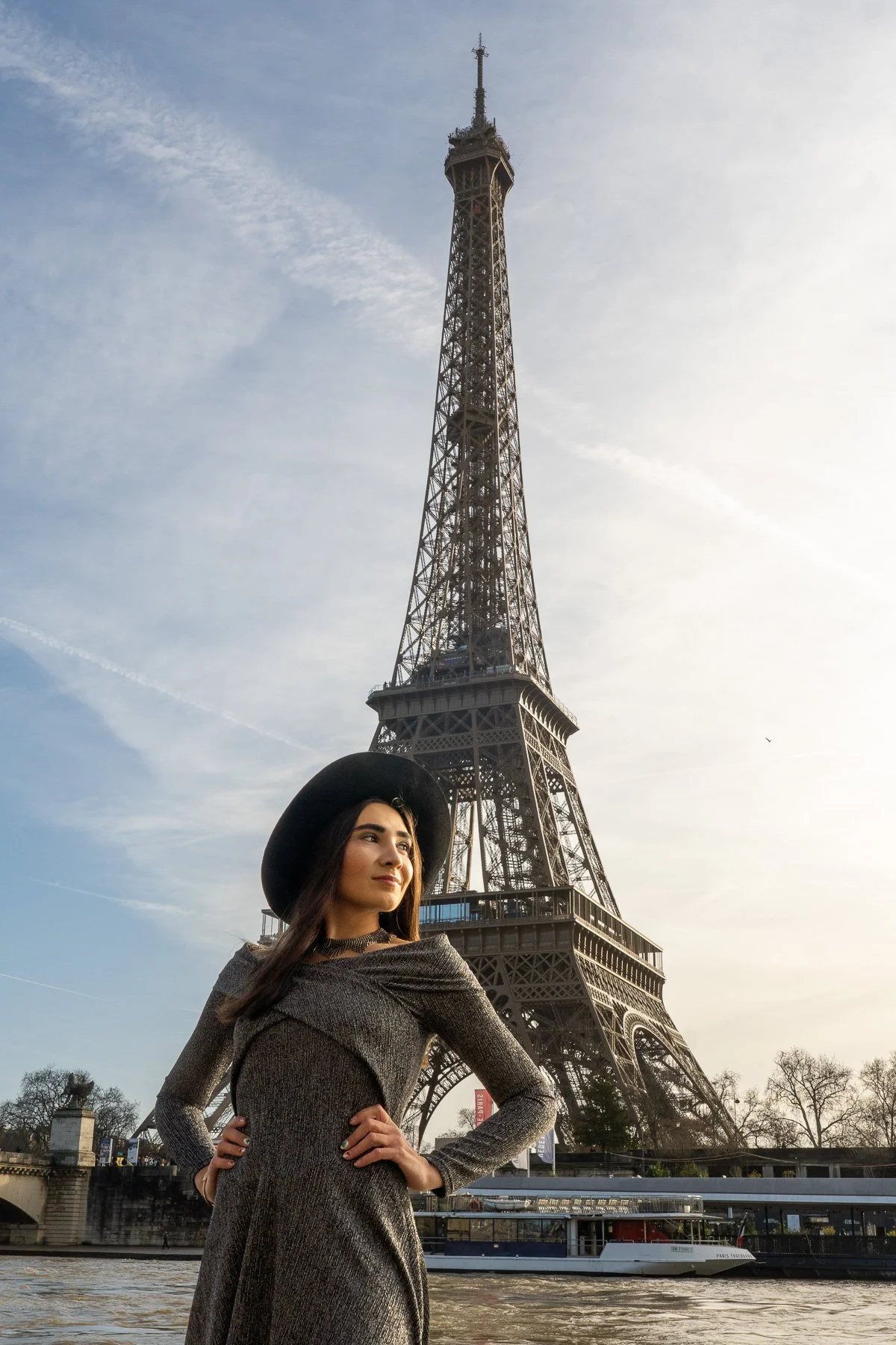 A woman in a gray dress and black wide-brim hat posing by the Seine River with the Eiffel Tower in the background during daytime.