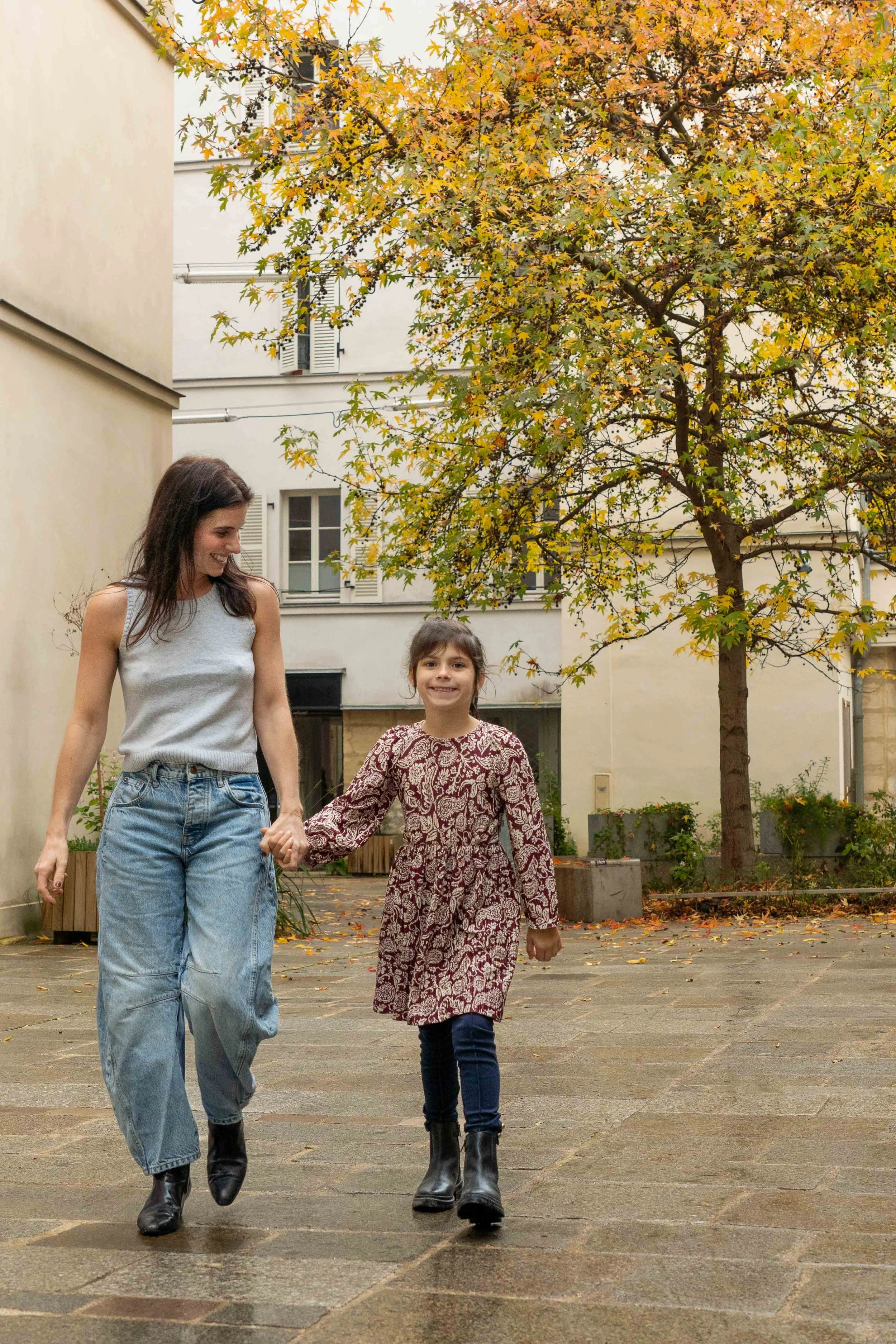 A woman and a young girl walk hand in hand outdoors on a paved surface, with trees and buildings in the background, during autumn.
