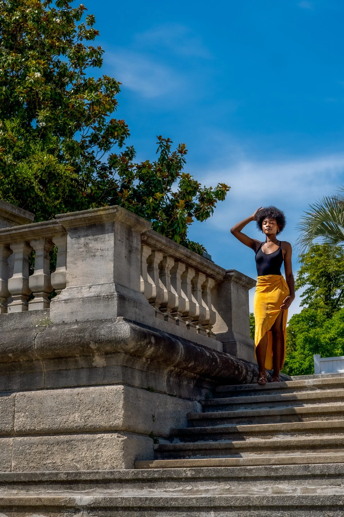 A woman standing on stone stairs outdoors, wearing a black sleeveless top and a yellow skirt with a slit, with green trees and a blue sky in the background.