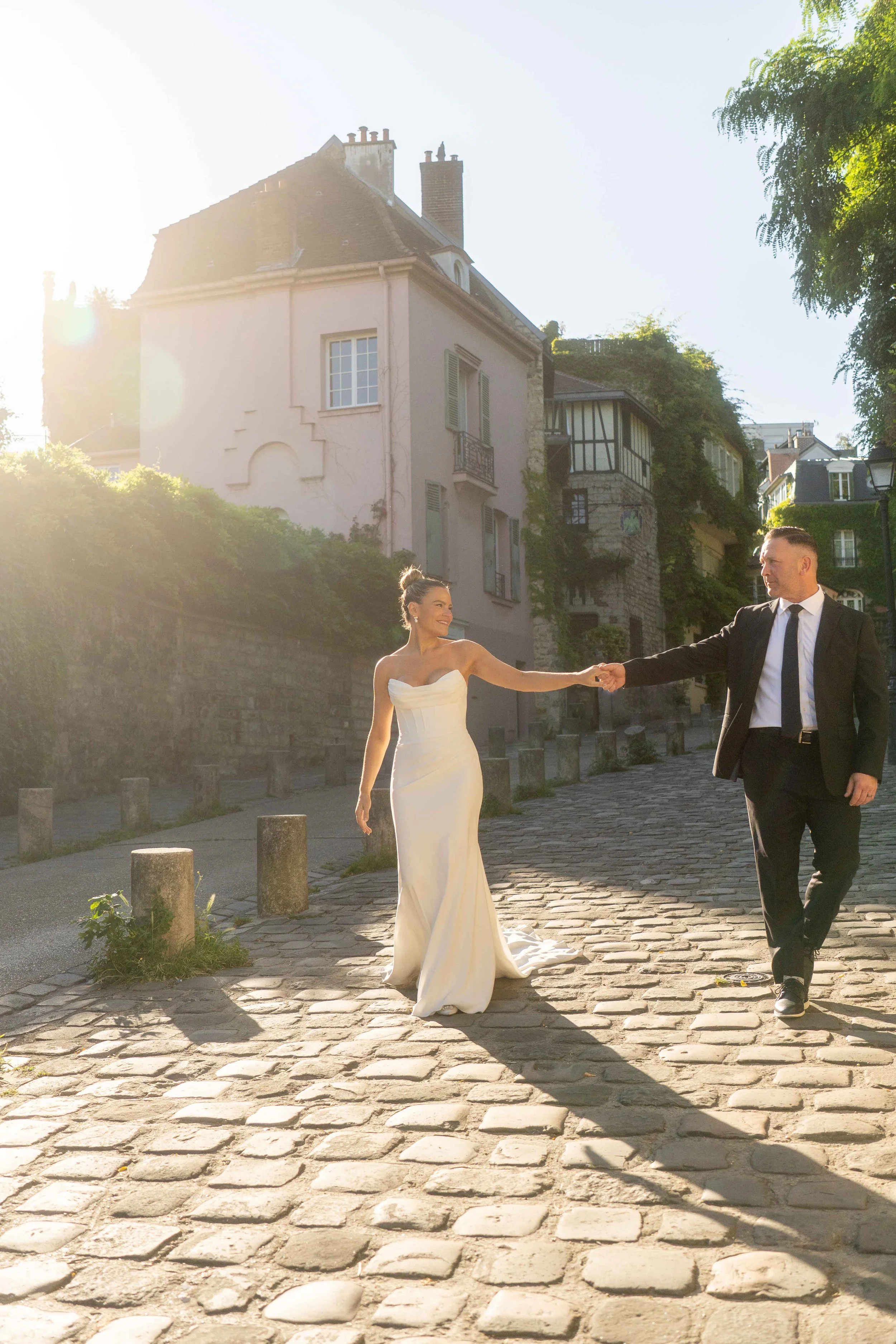 A bride and groom hold hands and walk on a cobblestone street in a European town, with the sun shining behind them. The bride is in a white wedding gown, and the groom in a black suit and tie.