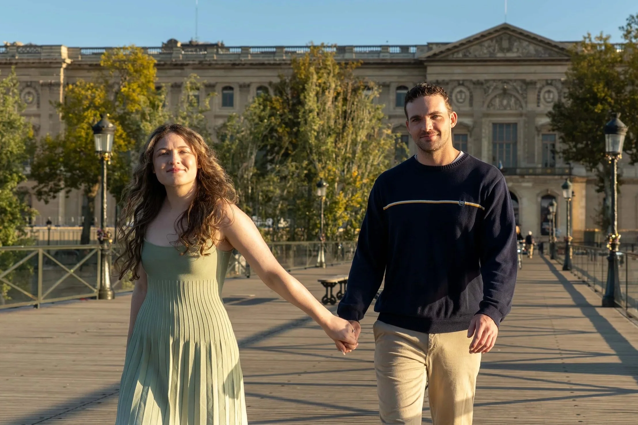 A young woman and man holding hands walking on a bridge in front of an ornate building during the daytime.