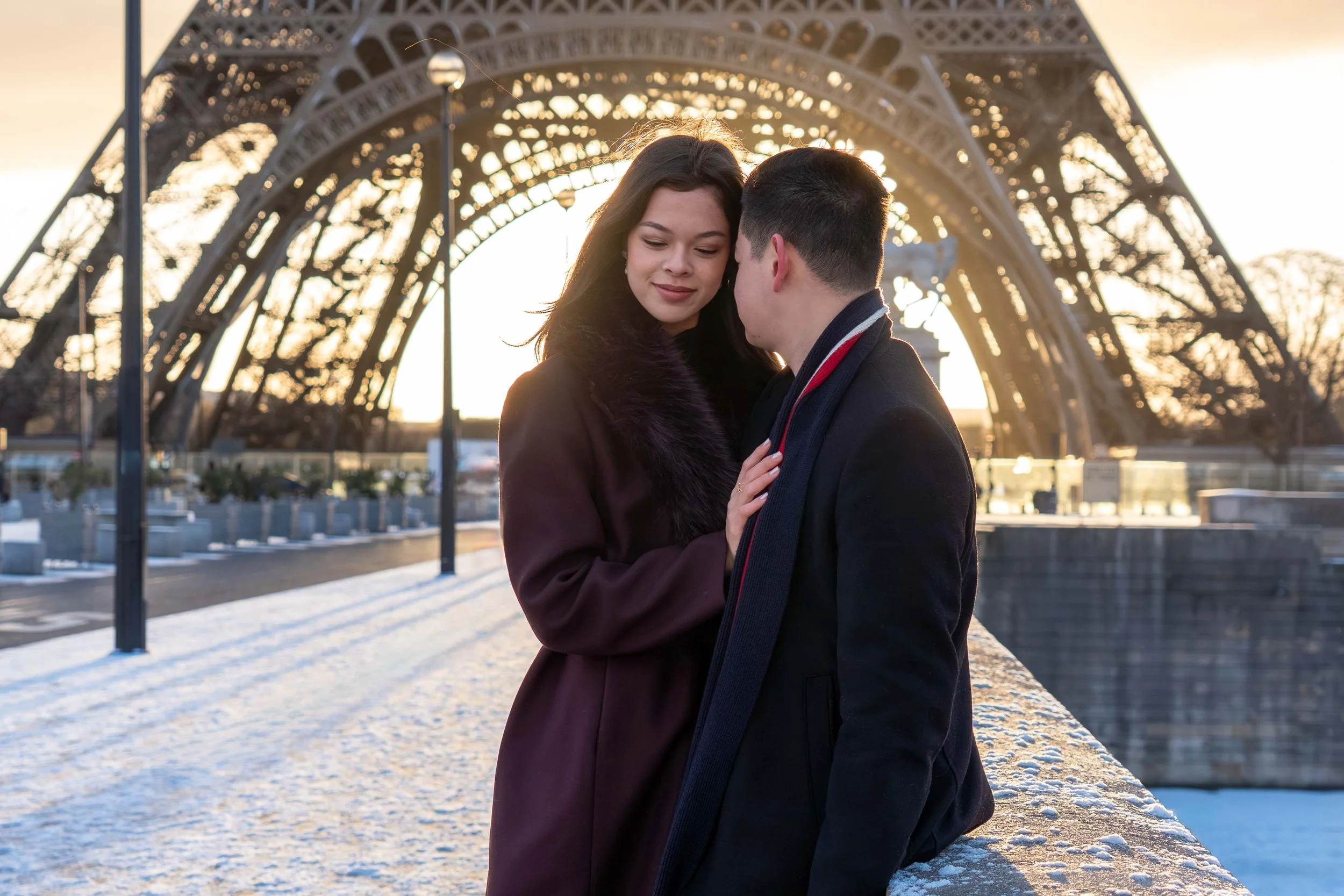 A couple close together on a bridge, with the Eiffel Tower in the background during sunset. The woman has dark hair, wearing a dark coat, and the man has short dark hair, wearing a dark coat and scarf. Snow is on the ground.