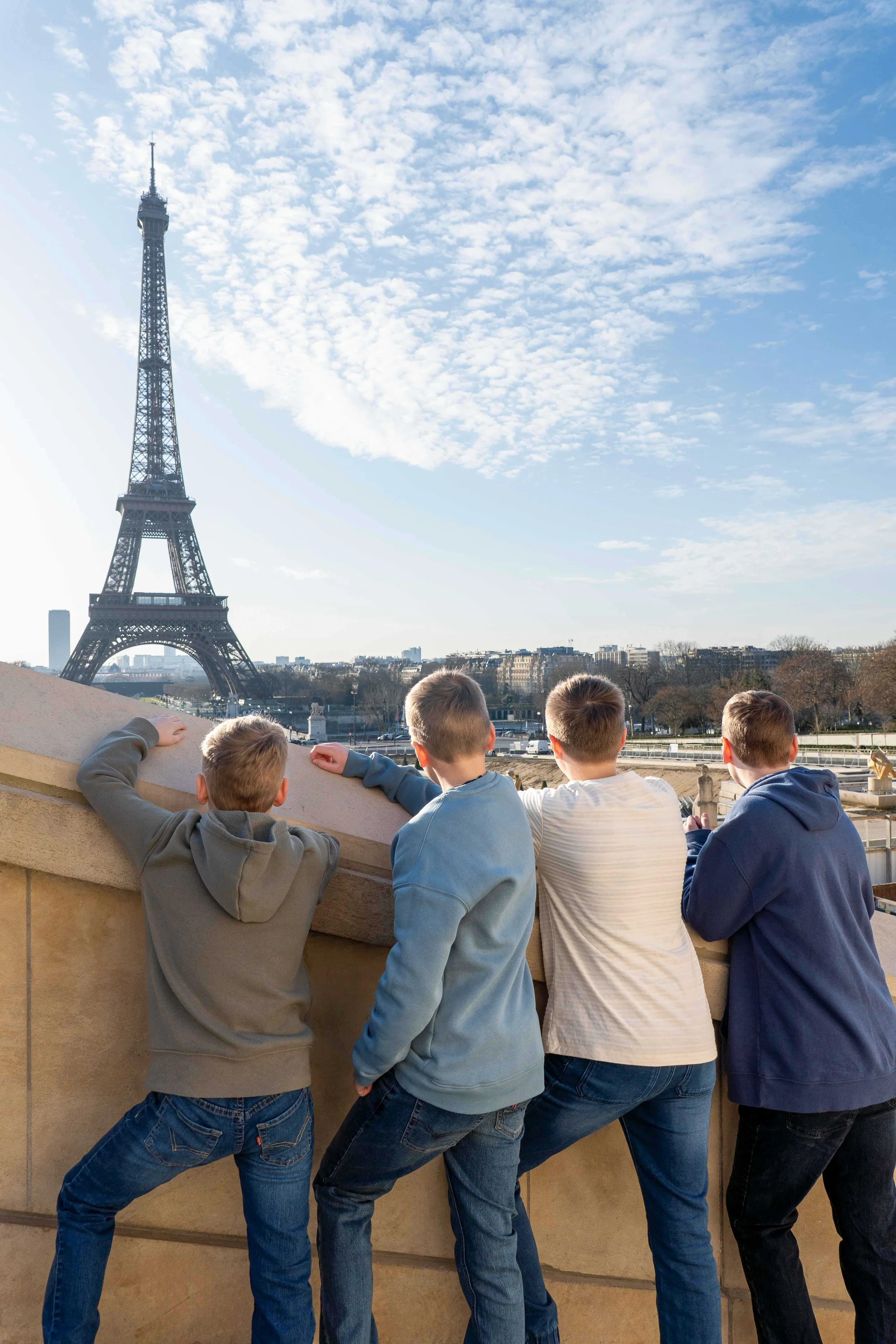 Four boys in casual clothing leaning on a stone railing, facing away, looking towards the Eiffel Tower and a blue sky with scattered clouds.