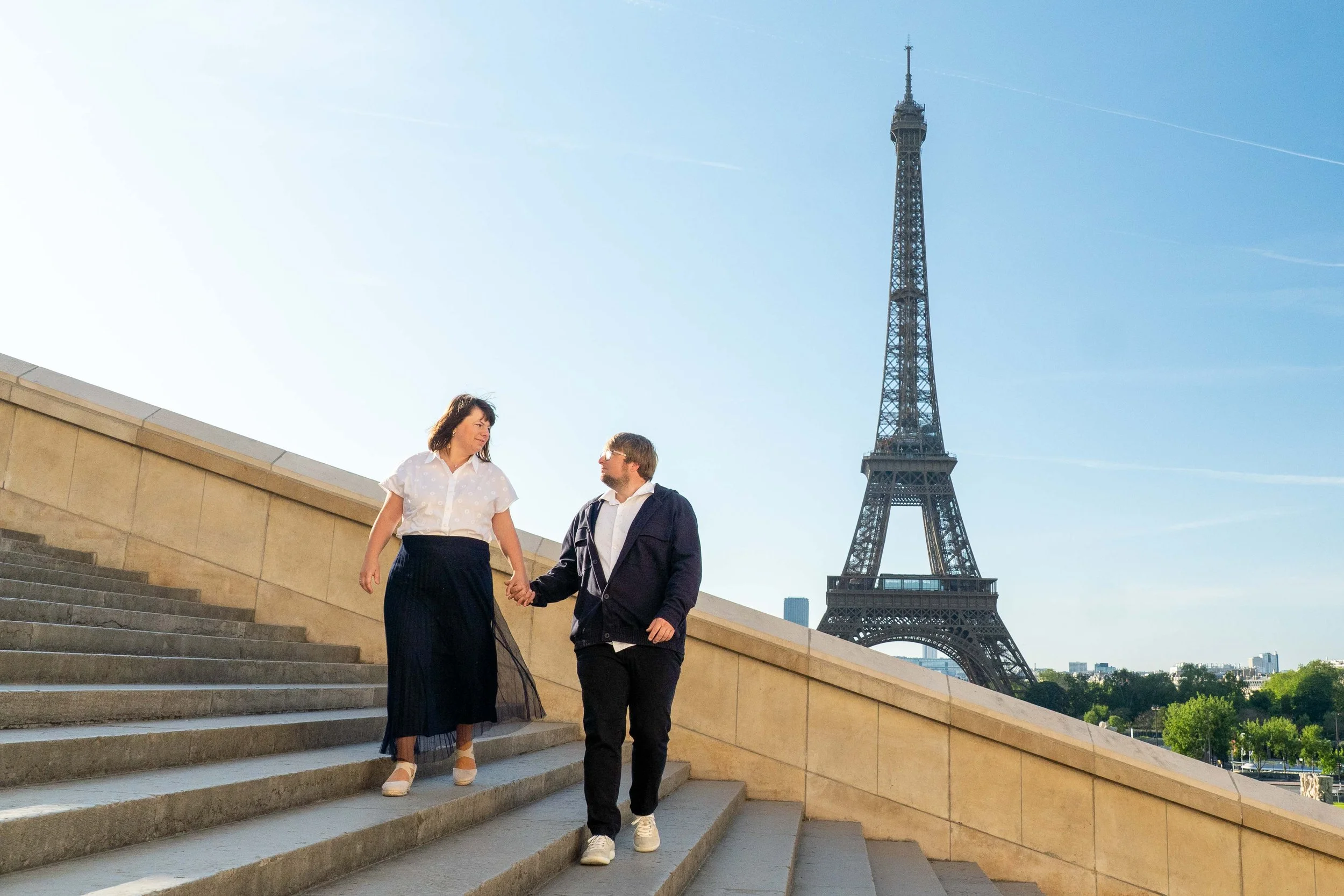 A couple holding hands walking down stairs near the Eiffel Tower in Paris, France, on a clear sunny day.