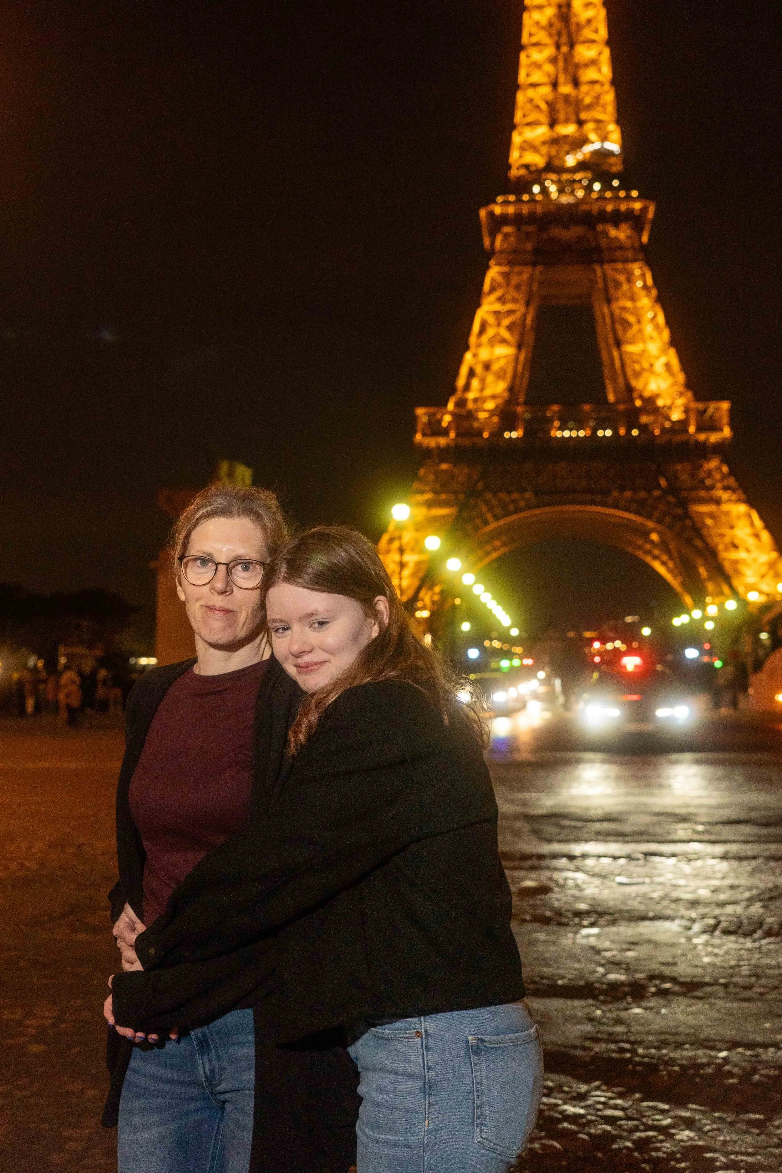 Two women hugging at night in front of the illuminated Eiffel Tower in Paris, France.