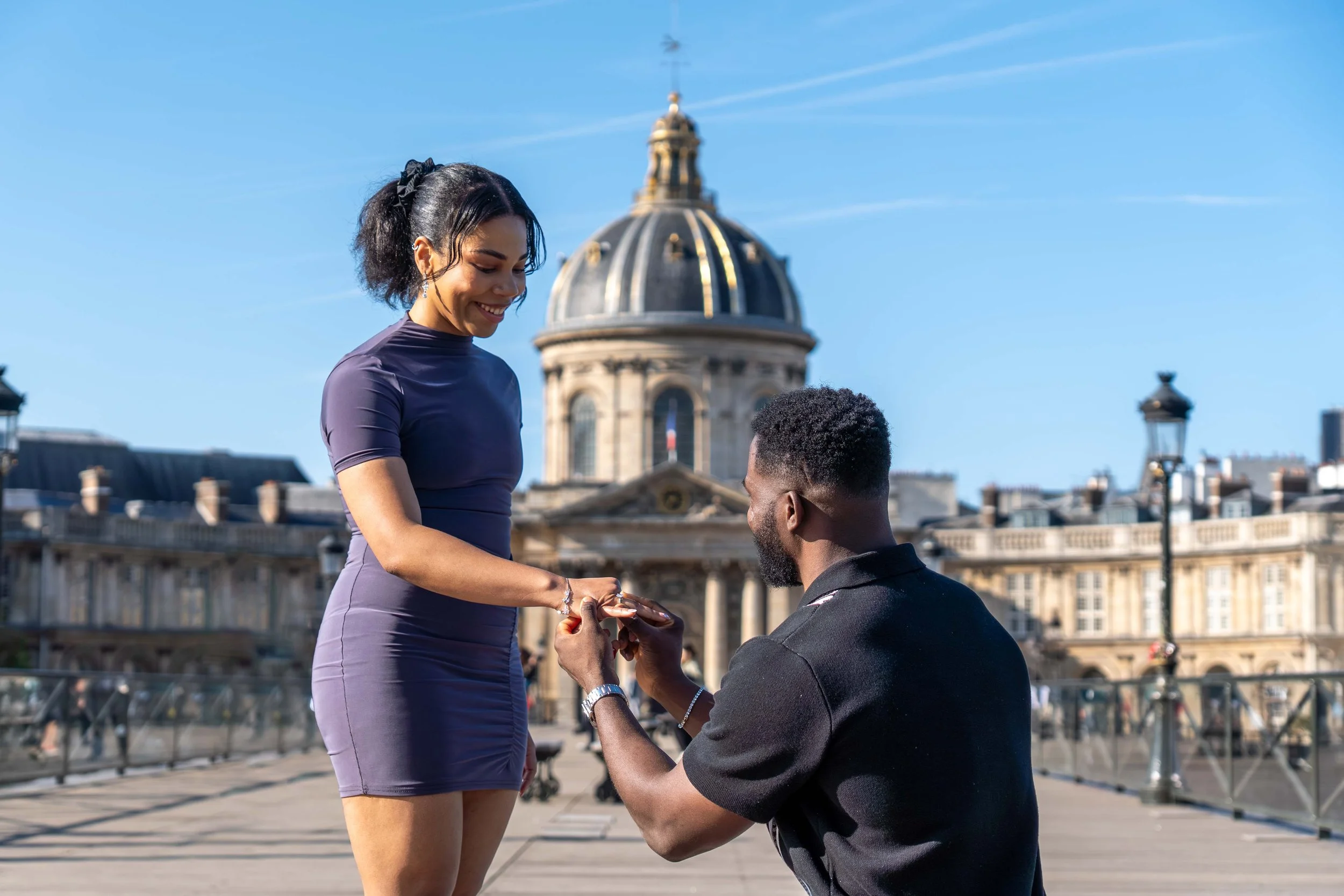 A man proposes to a woman on a city bridge with historic architecture and a dome in the background, under a clear blue sky.