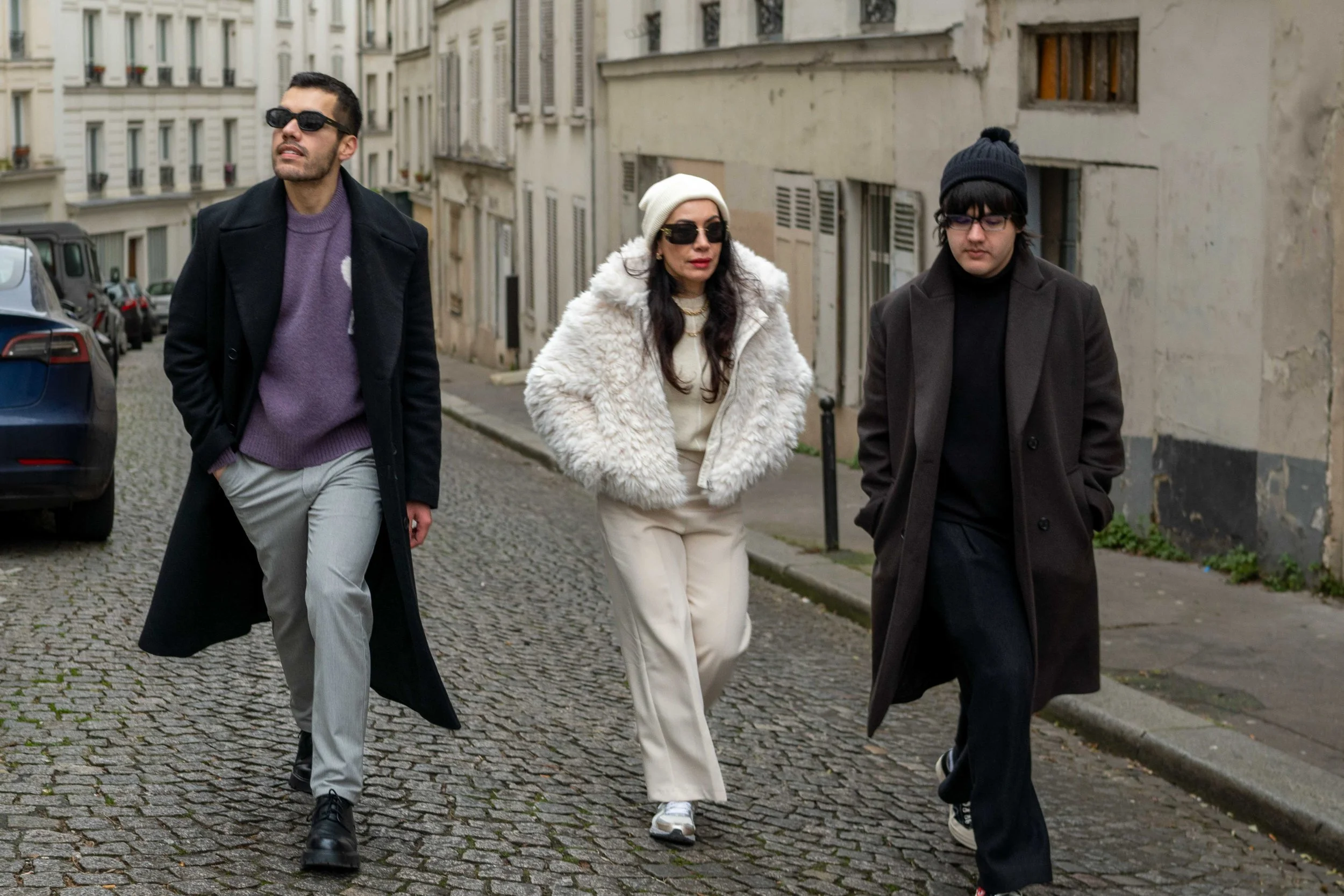 Three individuals walking on a cobblestone street in an urban area, dressed in stylish winter clothing, wearing sunglasses.