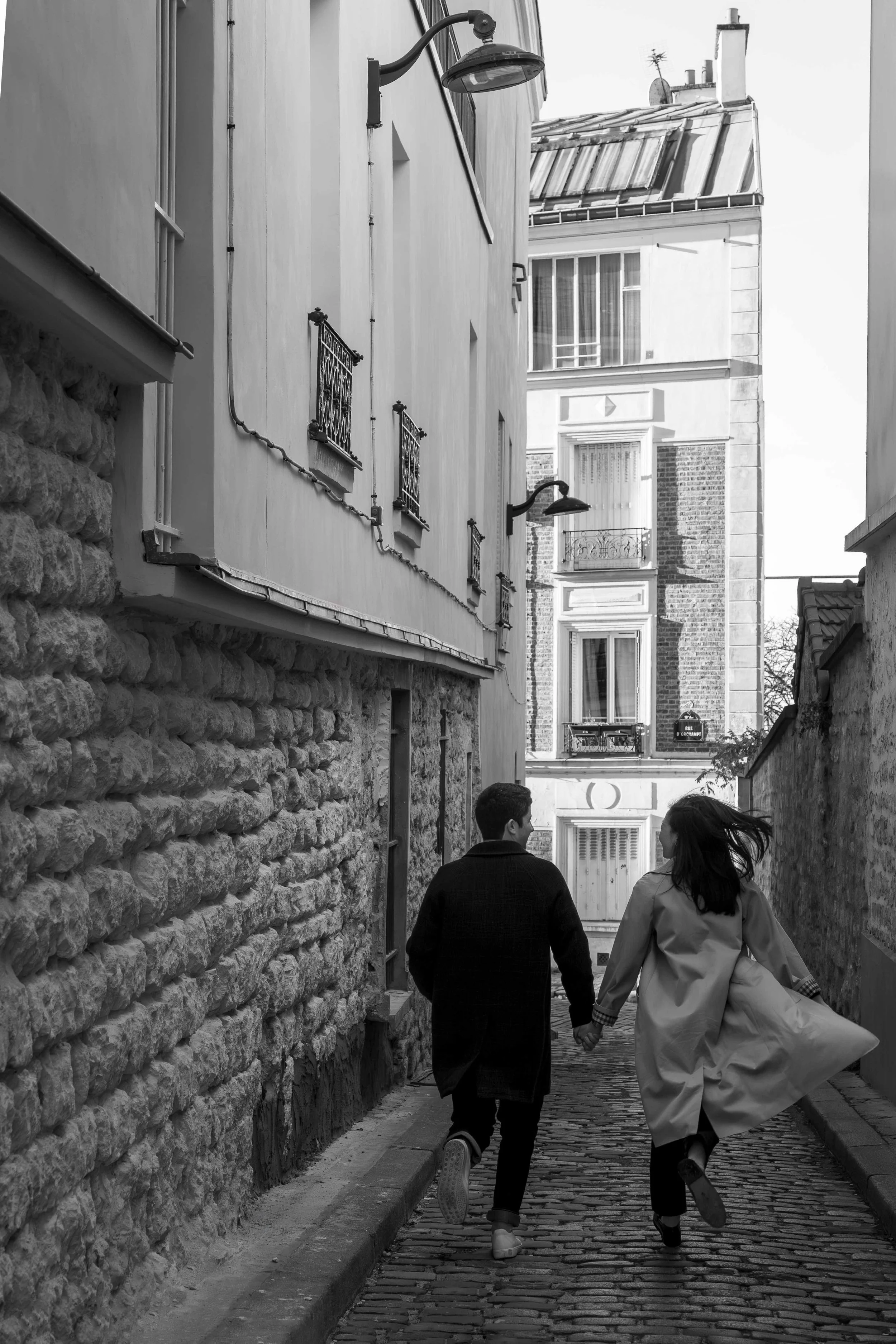 A black and white photo of a young couple holding hands and running down a narrow cobblestone street between old buildings in an urban setting.