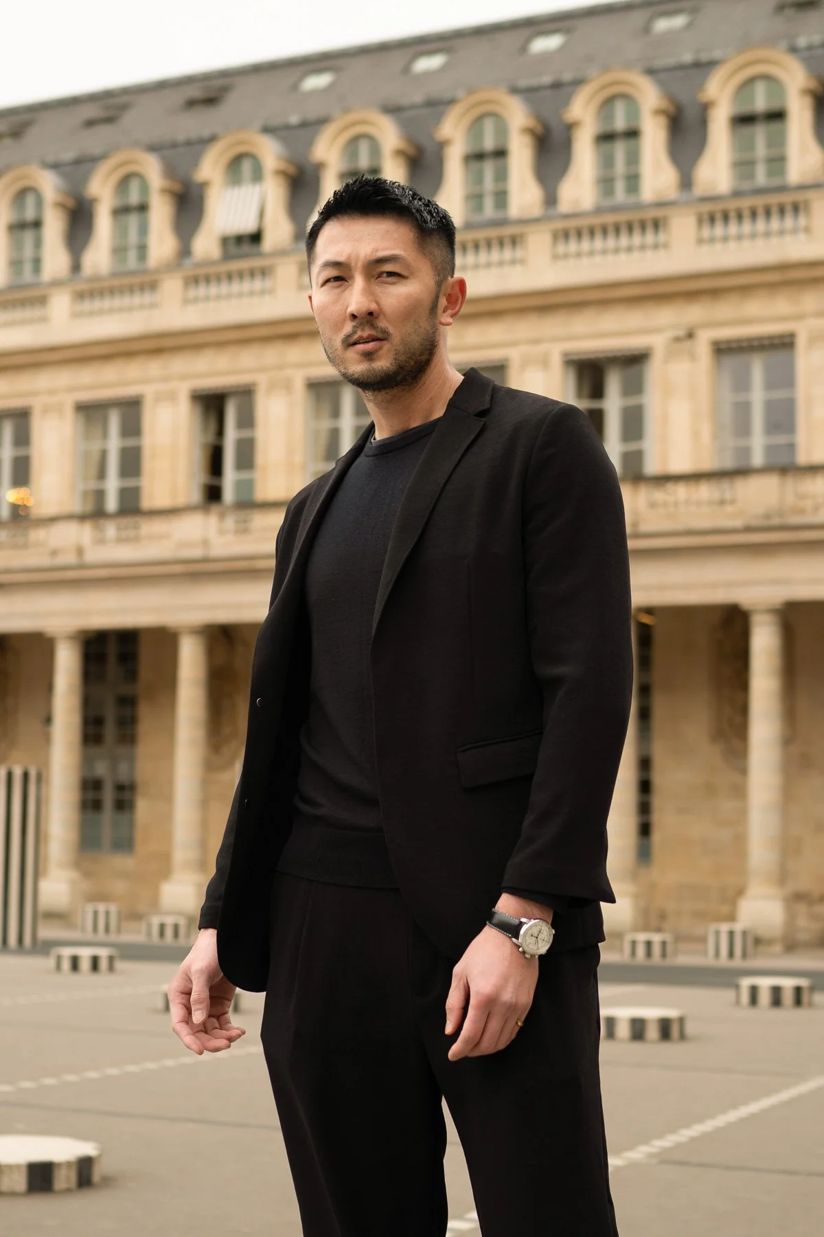 A man in a black suit standing outdoors in front of a historic building with large windows and ornate architectural details.