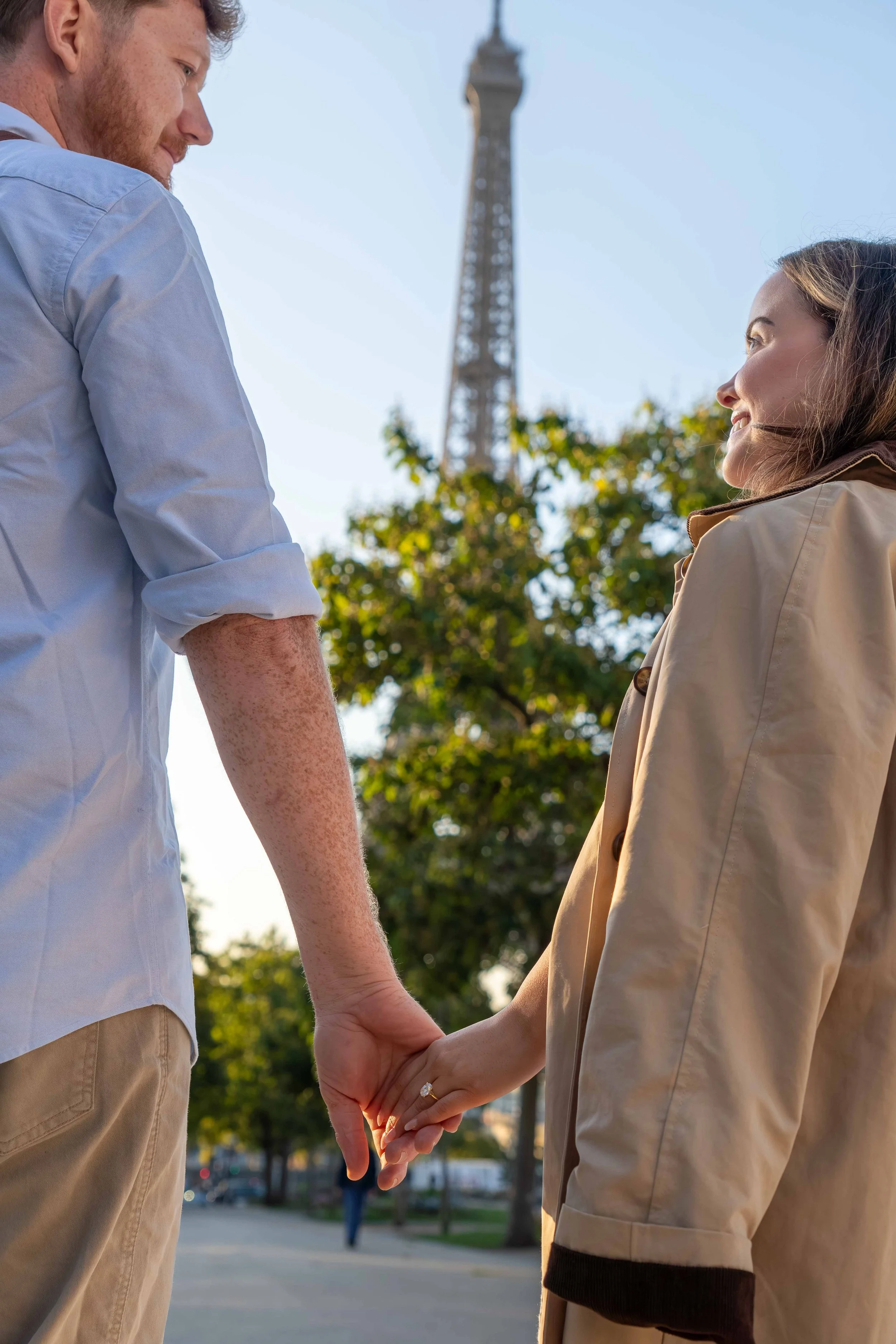 A man and woman holding hands outdoors near the Eiffel Tower, smiling at each other on a sunny day.