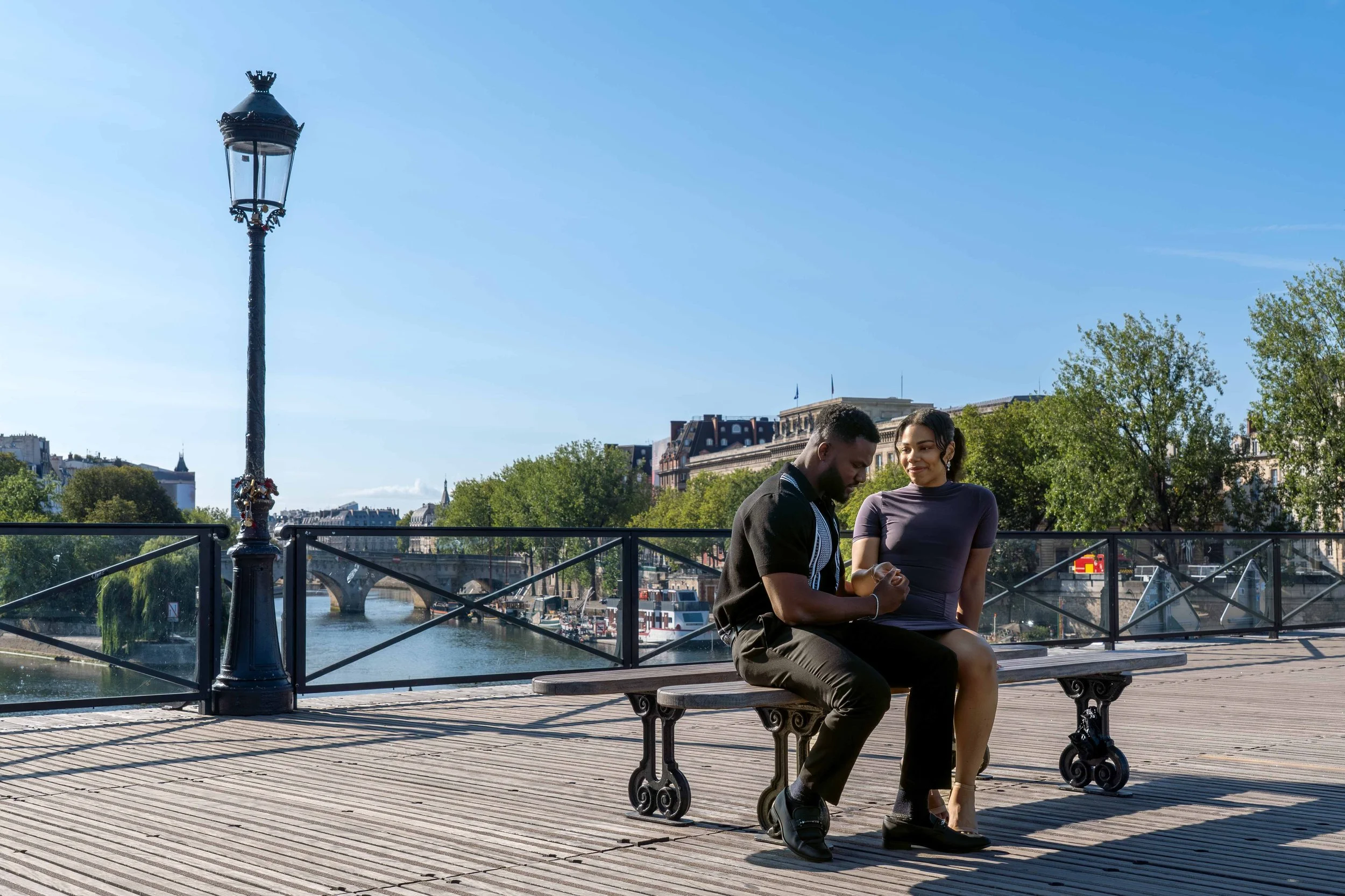 A couple sitting on a wooden bench by a river with bridge and city buildings in the background. The man is looking down at his hands, and the woman is smiling at him. There is a street lamp nearby.