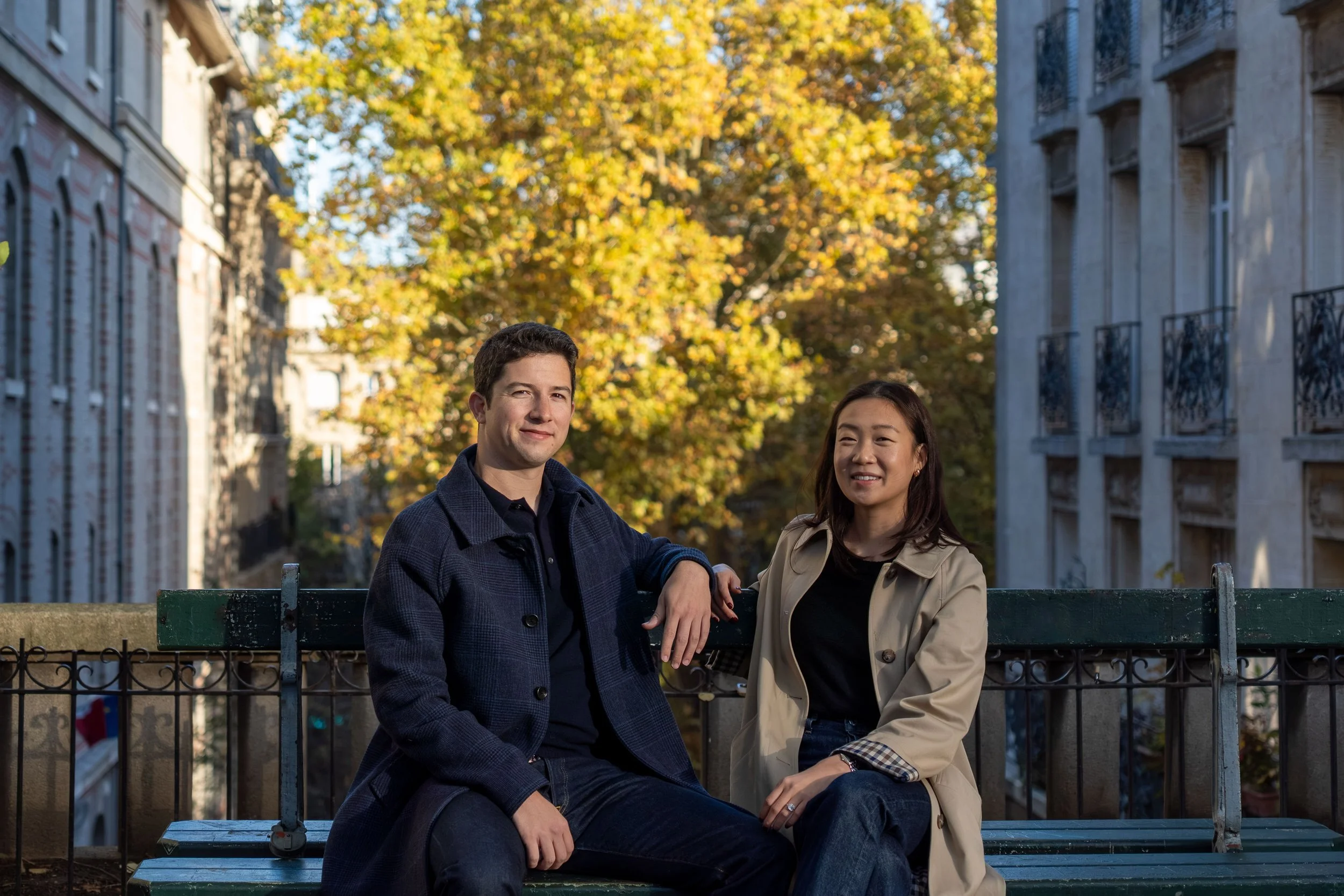 A man and woman sitting on a park bench outdoors during fall, with trees and buildings in the background.