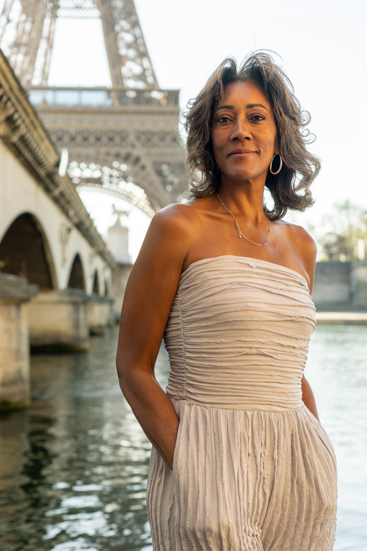 A woman in a strapless beige dress standing by the Seine River in Paris with the Eiffel Tower in the background.