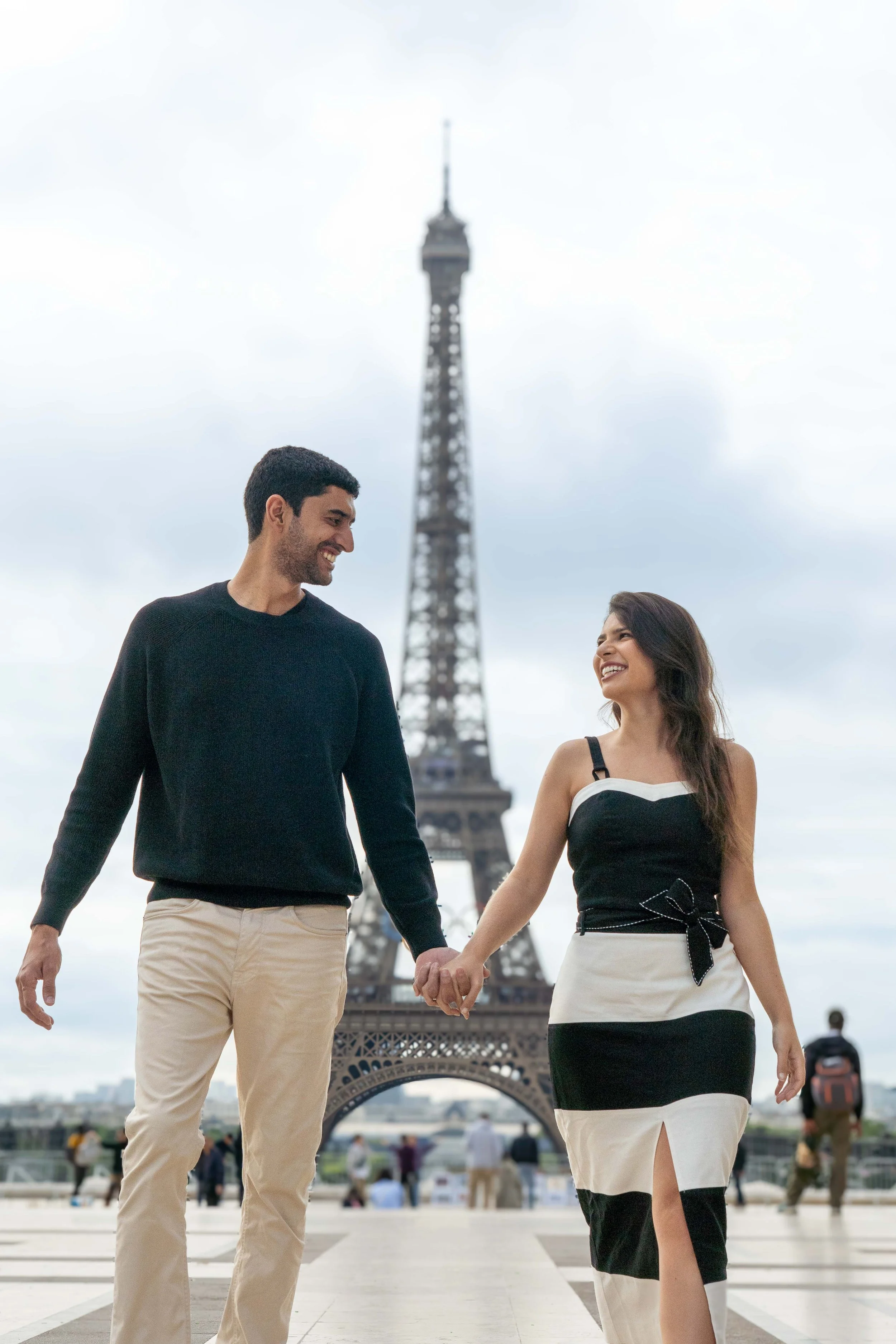A man and woman holding hands and smiling at each other in front of the Eiffel Tower in Paris.